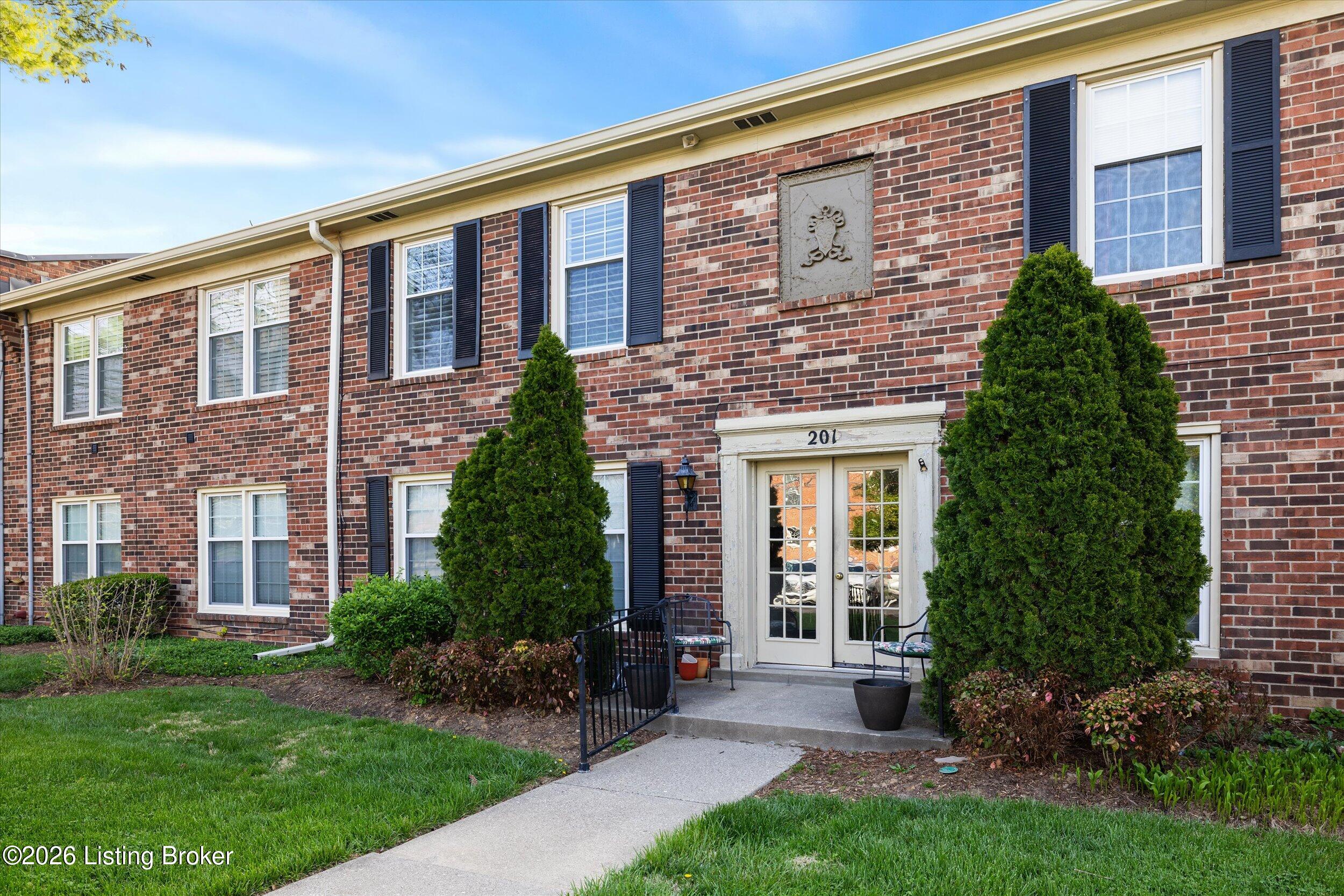 Brick residential building entrance with black shutters, landscaped shrubs, and glass-paneled doors marked 201.