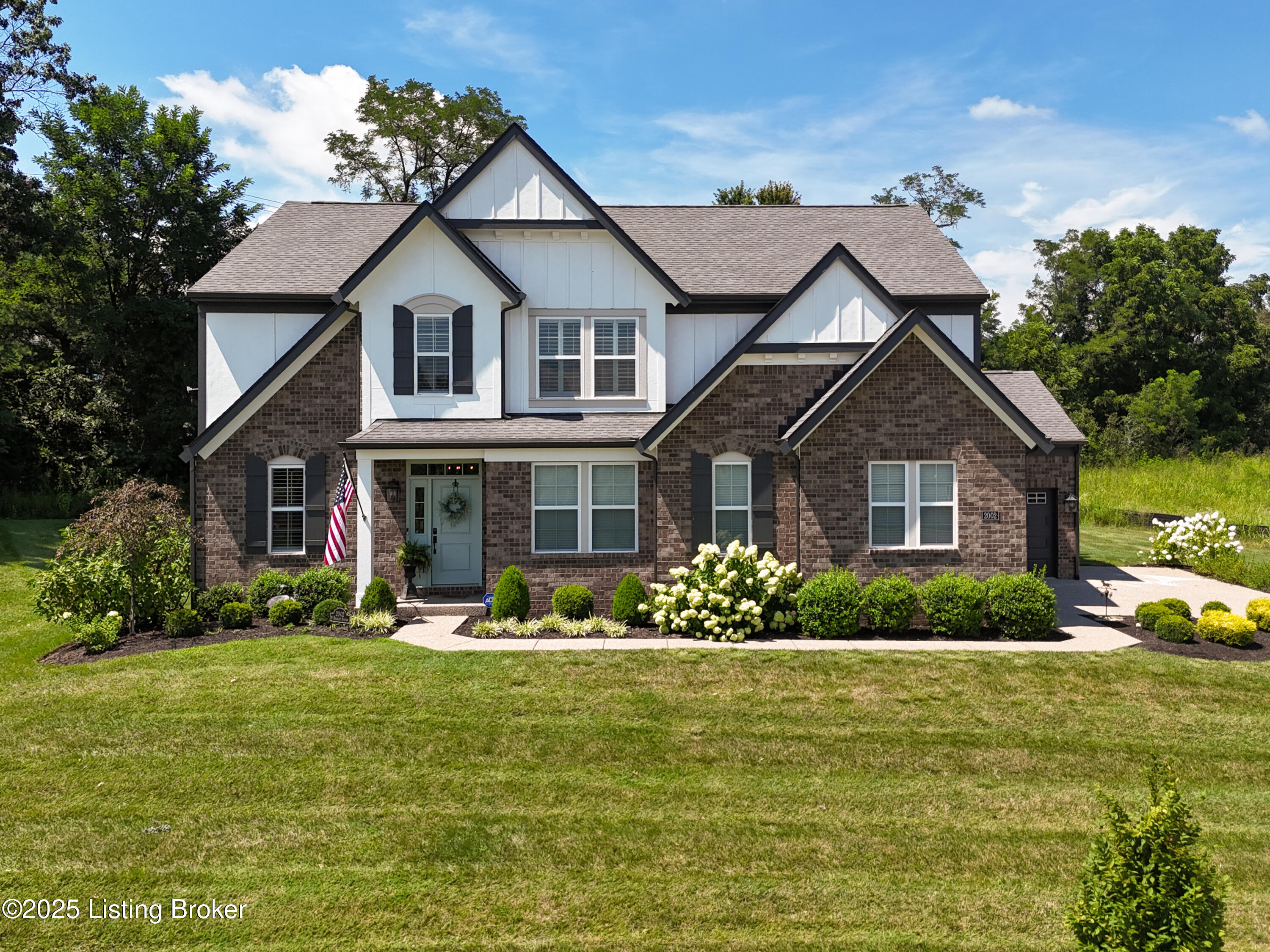 Two-story brick-and-white home with front gables, landscaped lawn, flowering shrubs, and an American flag.