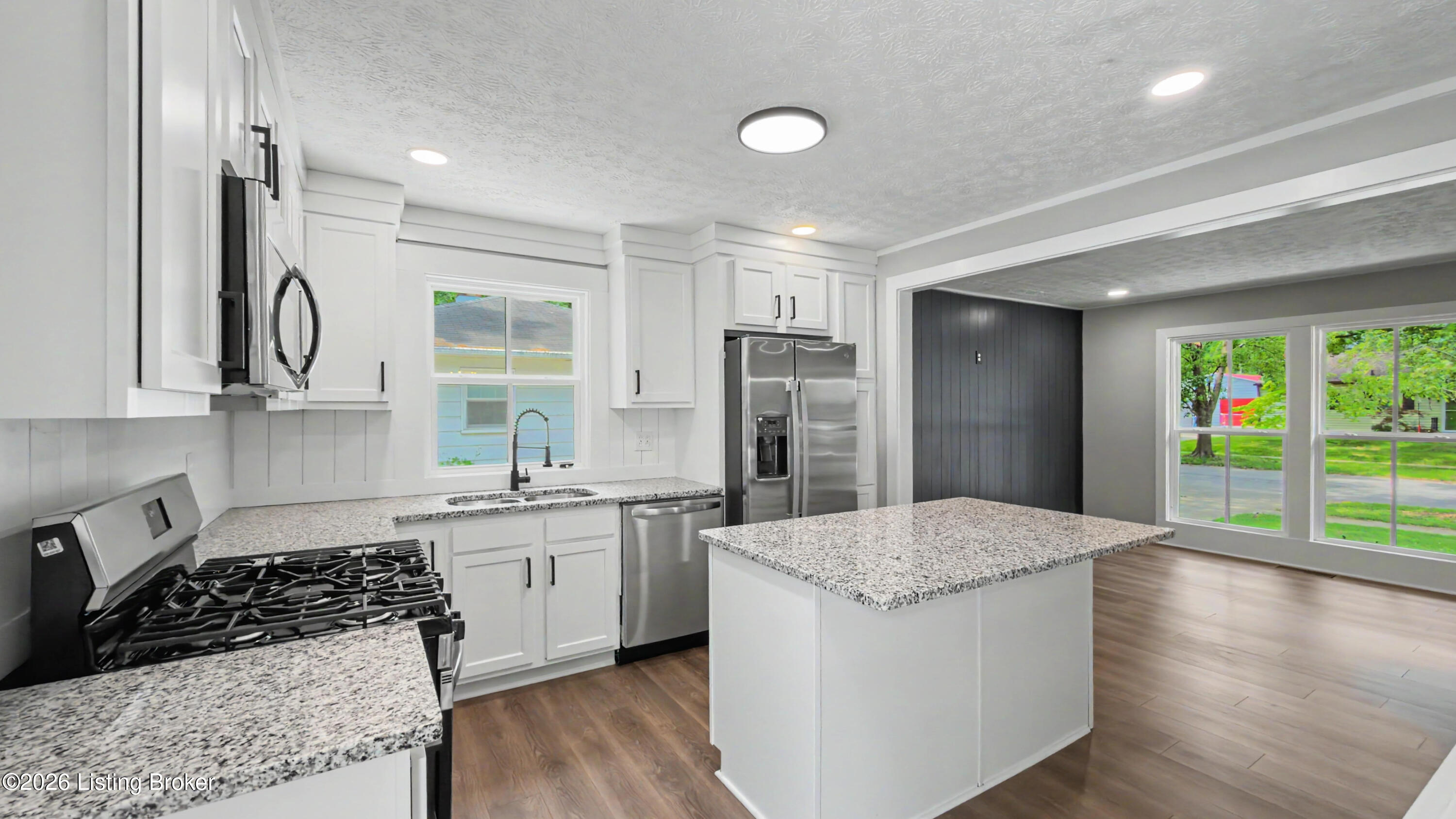 Renovated white kitchen with granite island, stainless appliances, and large windows opening to a living area.