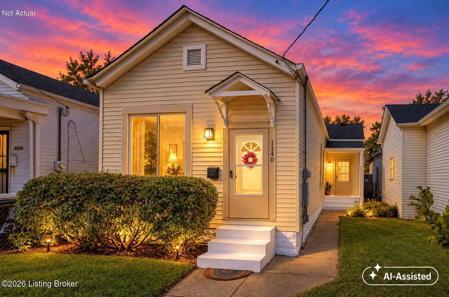 Small light-sided home with an illuminated entry and landscaped shrubs beneath a vivid sunset sky.