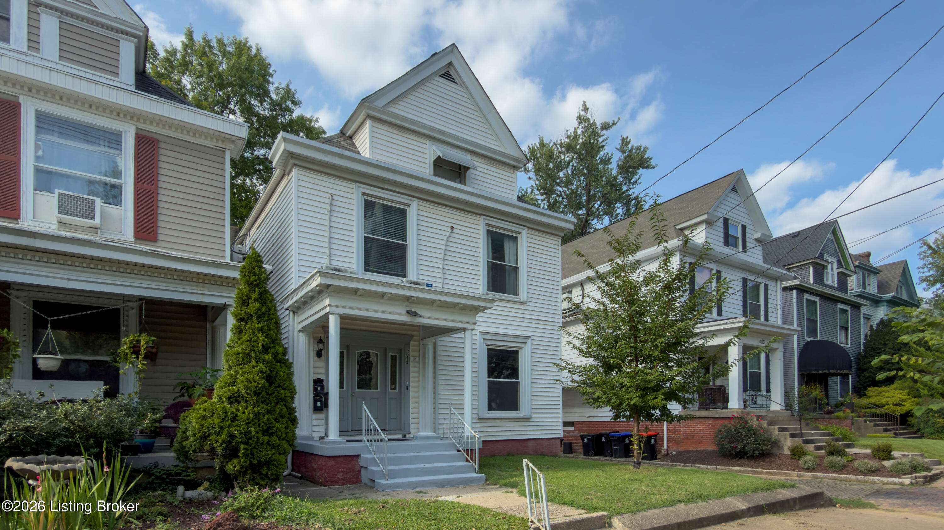 White clapboard house with a covered porch and front steps, flanked by neighboring homes.