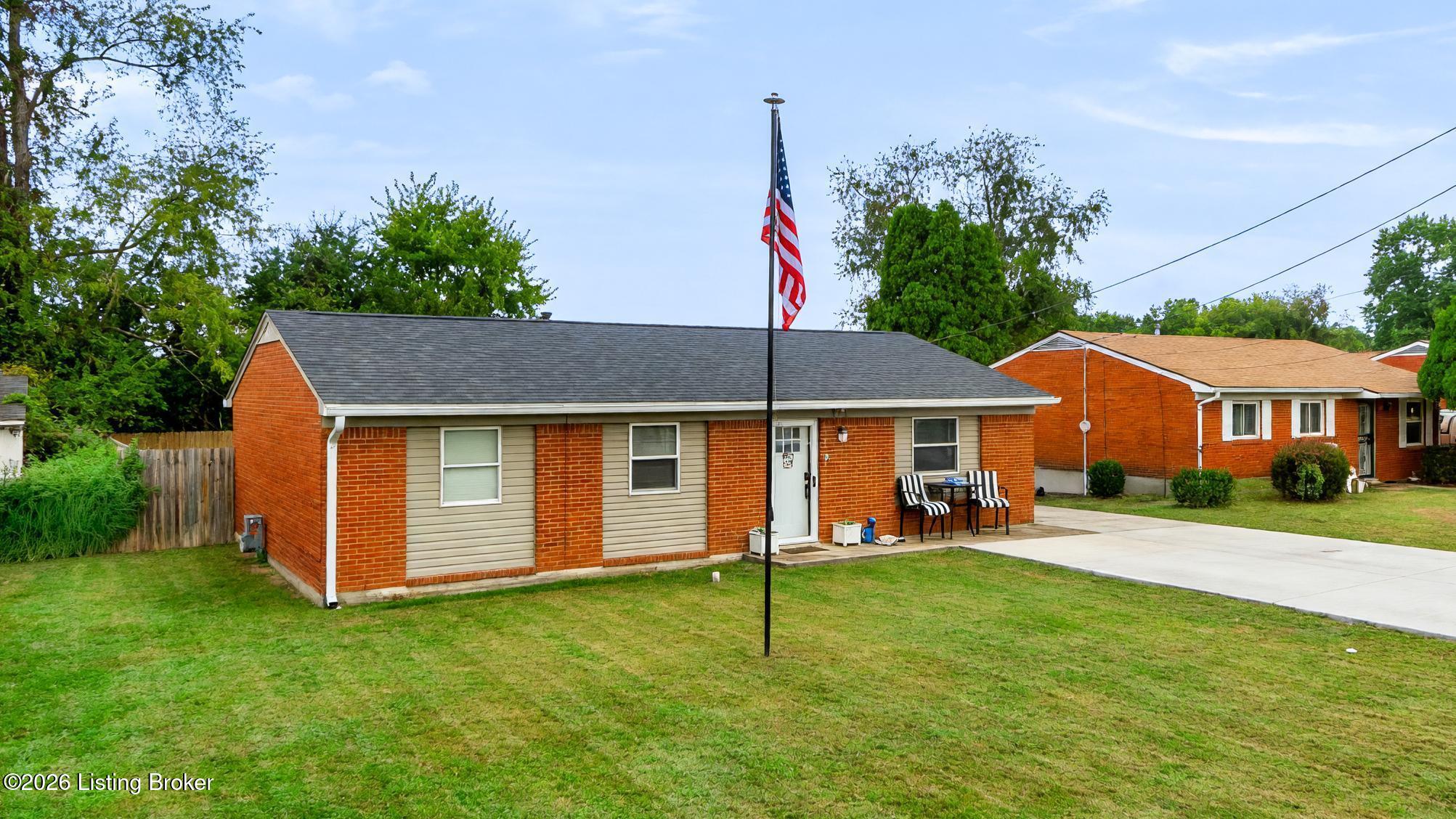 Single-story brick house with an American flag, striped patio chairs, and a broad front lawn.