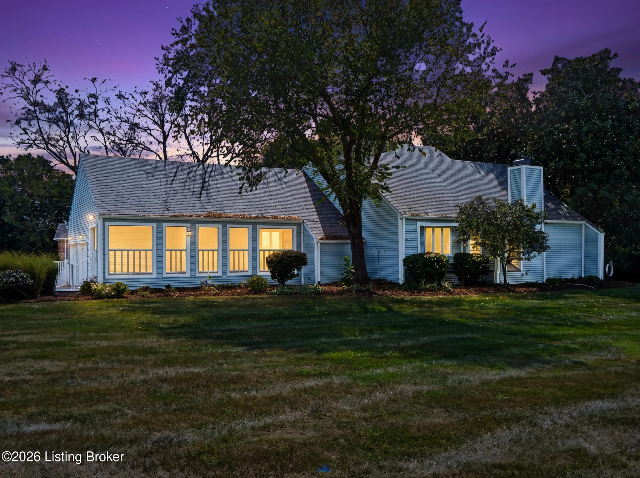 Blue-gray home with warmly lit windows beneath a large tree at dusk.