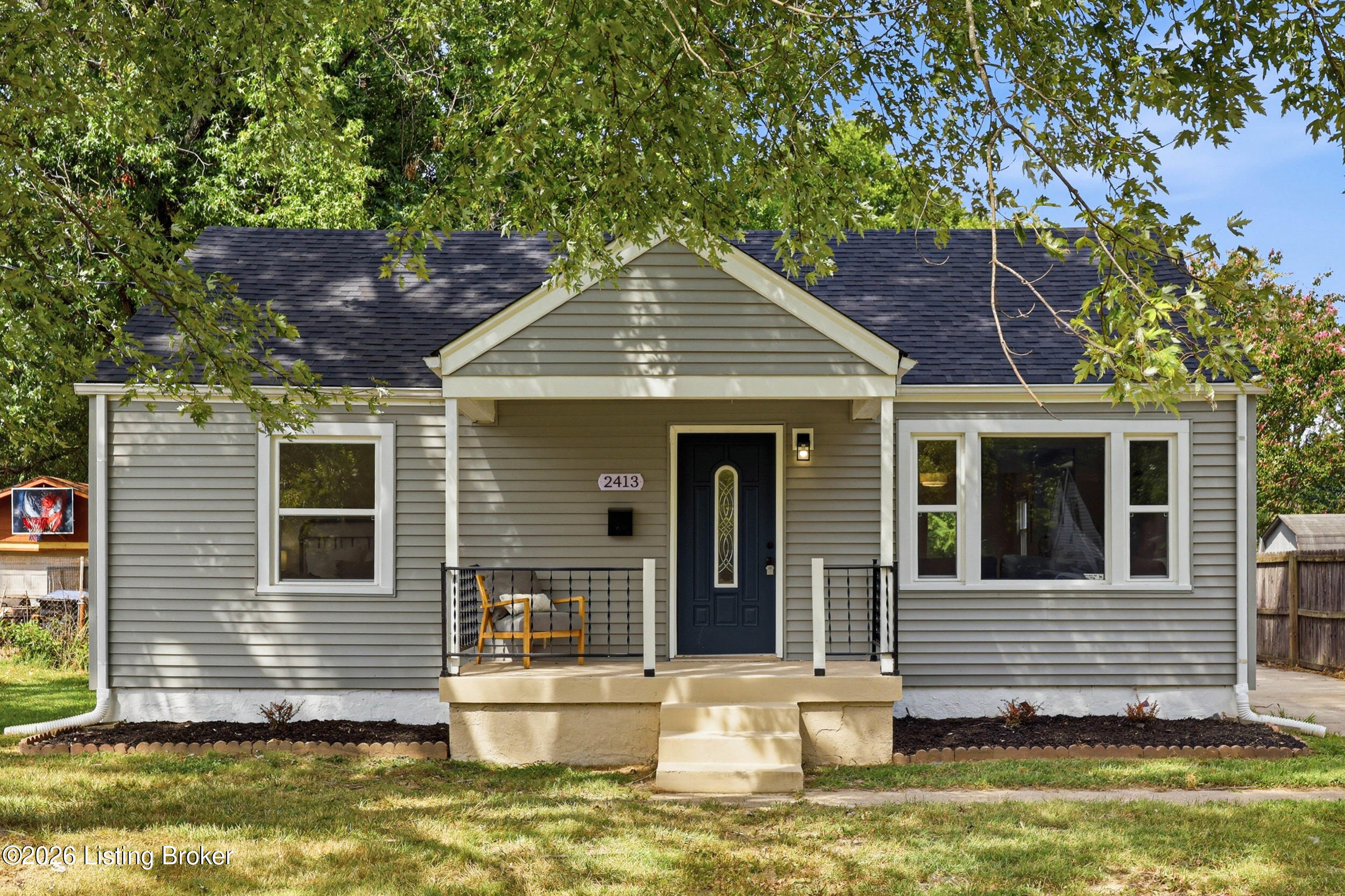 Gray single-story house with a covered porch, dark blue front door, and leafy tree overhead.