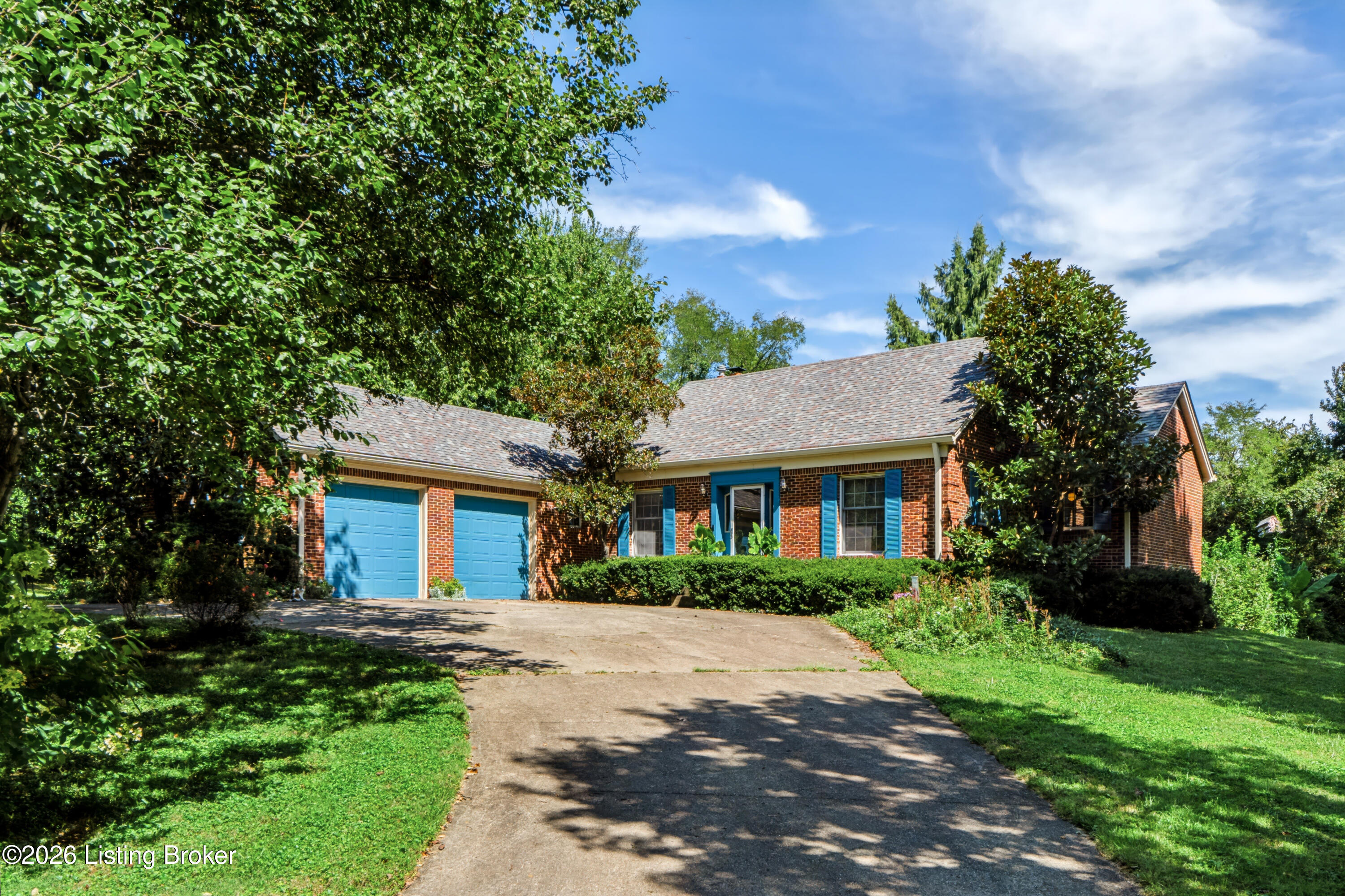 Red-brick house with blue garage doors and shutters, framed by mature trees and a broad driveway.