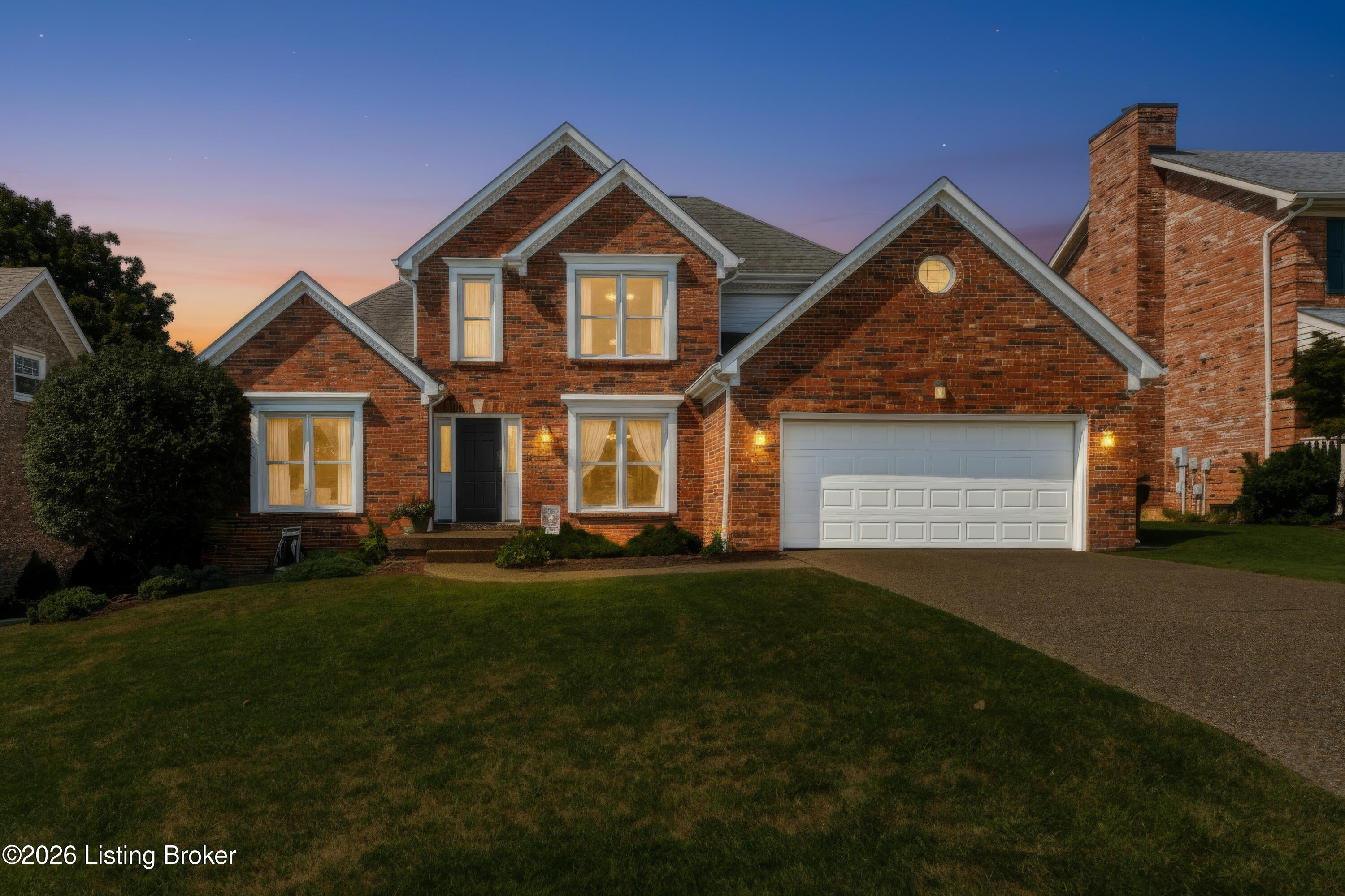 Two-story brick house with a lit circular window, white garage door, and landscaped lawn at dusk.