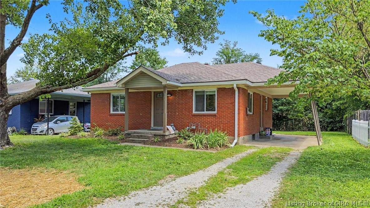 Red brick single-story home with a covered driveway, gravel path, and mature trees.