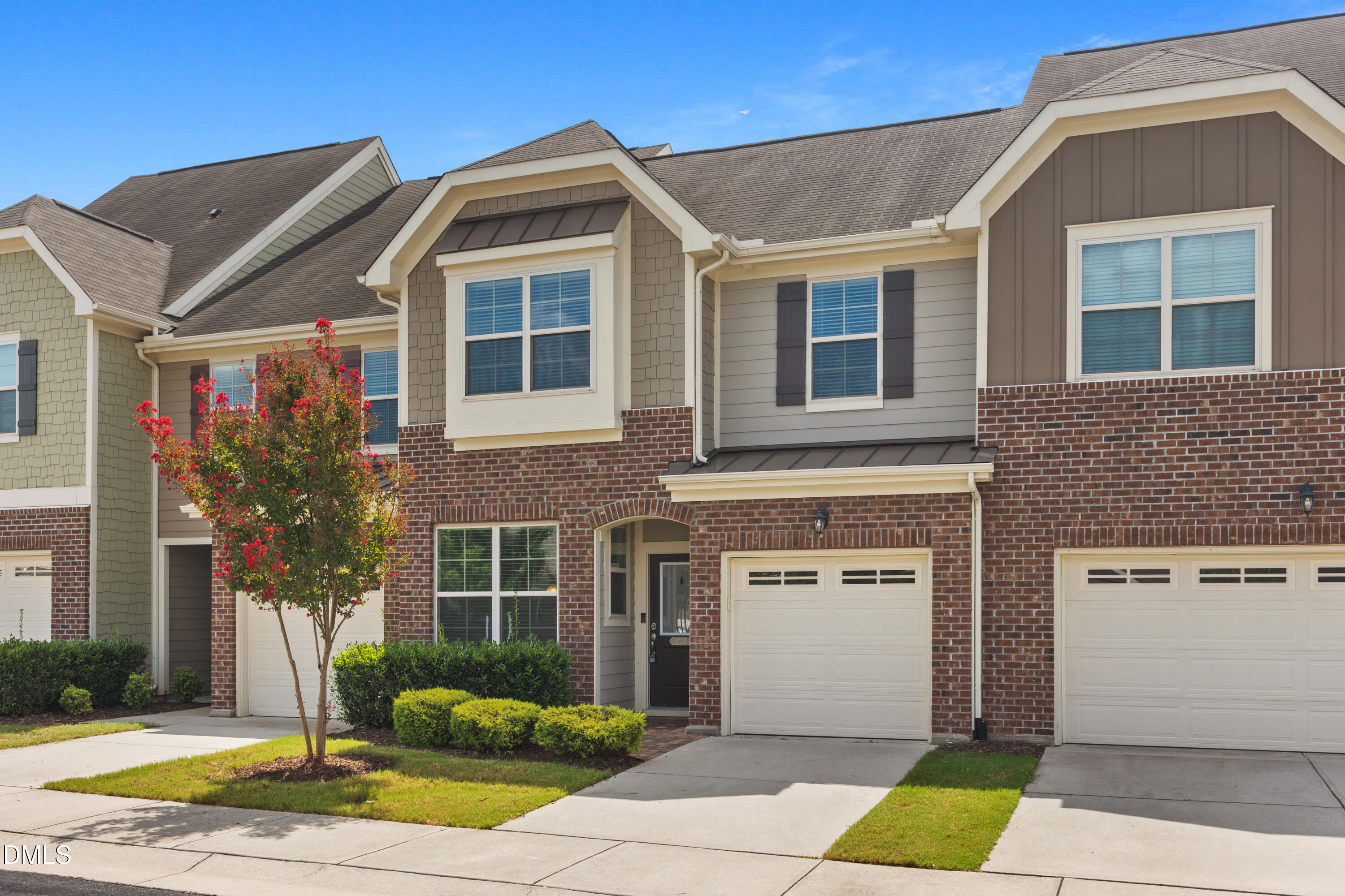 Attached townhomes with brick facades, white garage doors, and a flowering red tree.