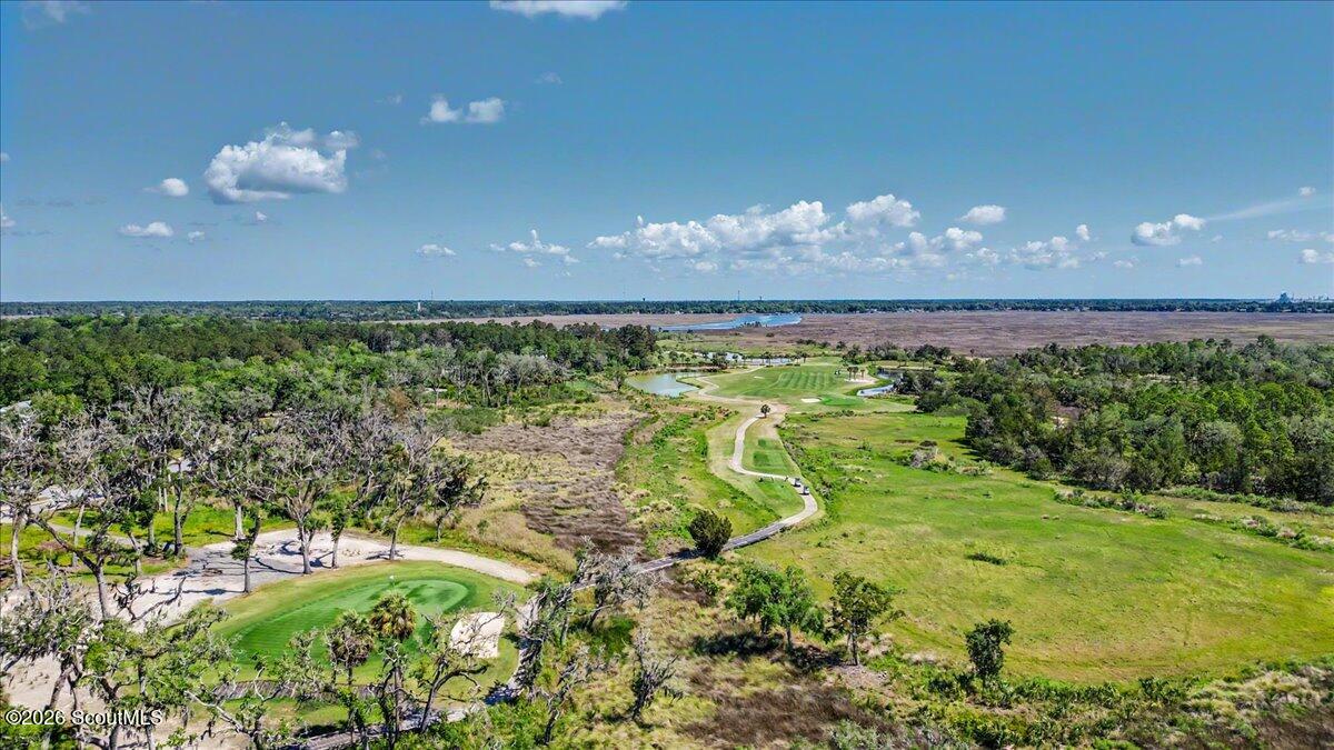 Aerial view of green golf-course grounds, a winding cart path, wooded areas, and distant marshland and water.