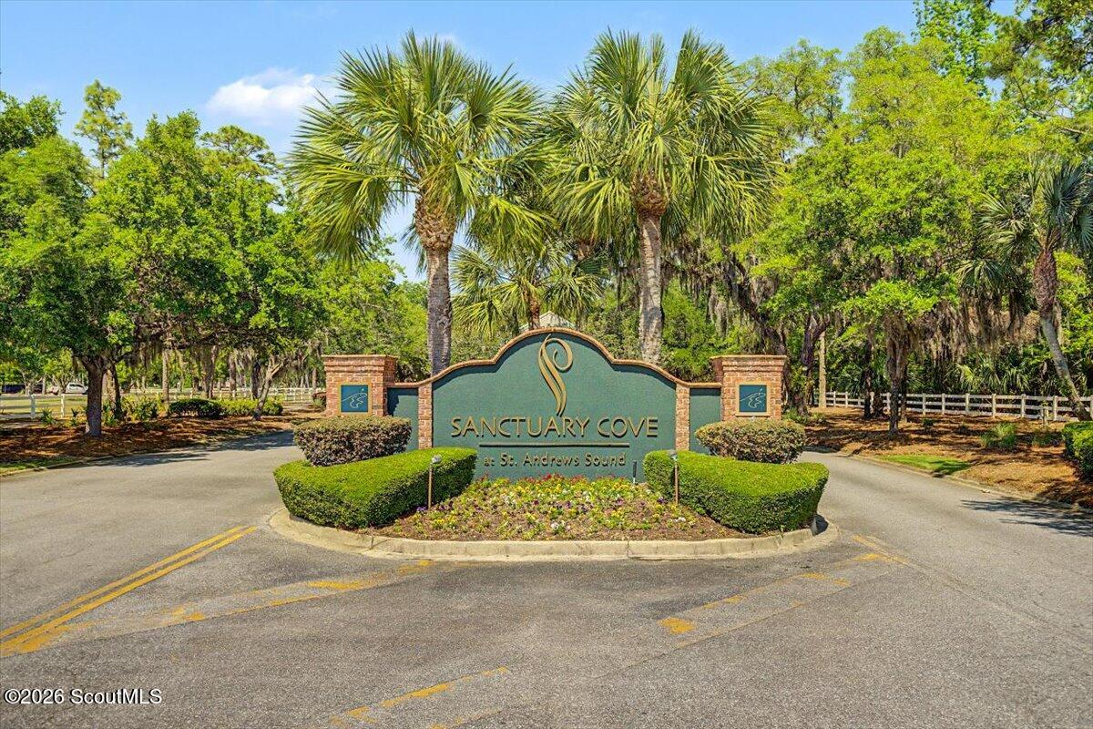 Sanctuary Cove entrance sign framed by palm trees and brick pillars beside a curving road.