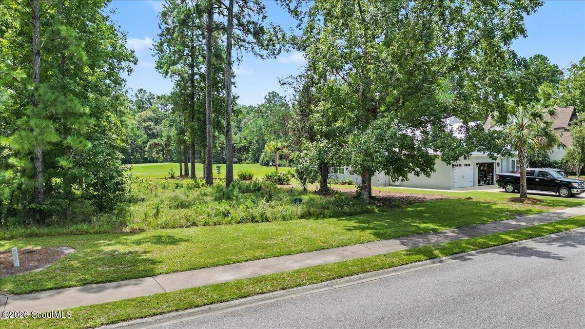 Vacant tree-lined lot beside a sidewalk, with a manicured golf course visible beyond it.