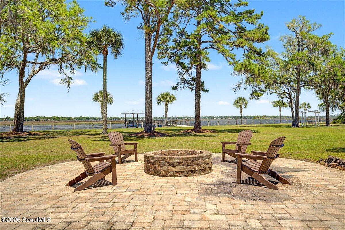 Waterfront patio with a circular stone fire pit, Adirondack chairs, mature trees, and a dock.