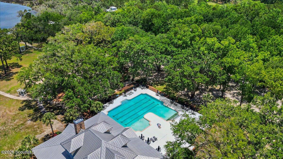 Aerial view of a swimming pool beside a gray-roofed building, surrounded by trees with a pond nearby.