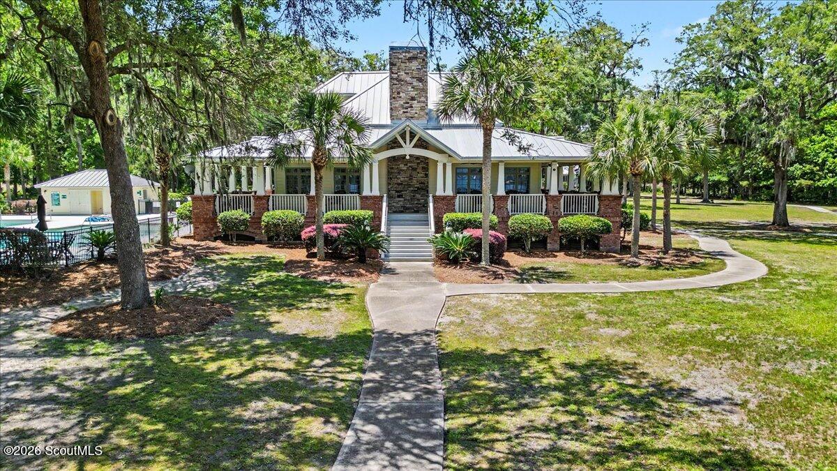 White building with a stone chimney, broad covered porch, palm trees, and a pool beside the lawn.