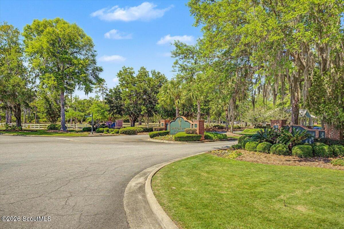 Neighborhood entrance with a curved road, landscaped lawn, and mature trees.