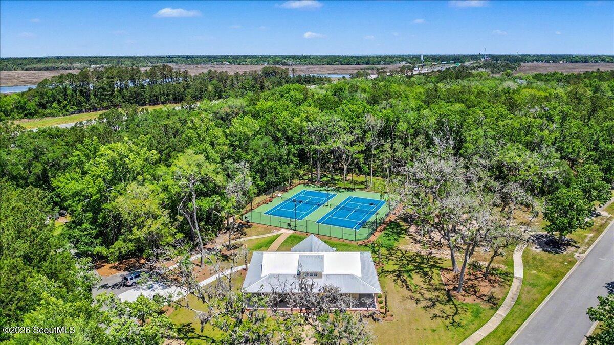 Aerial view of two tennis courts surrounded by trees, with a white-roofed building and distant marshland.