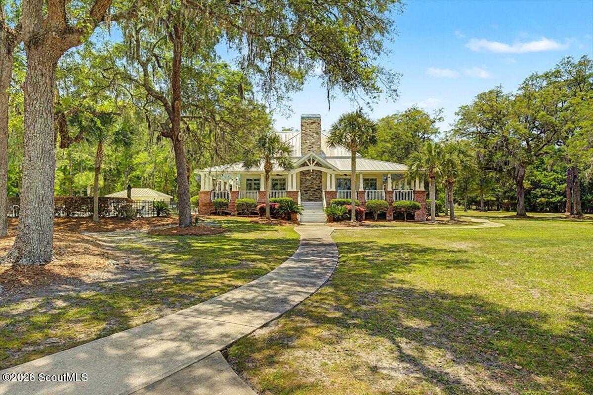 Low country-style building with a stone chimney and covered porch, surrounded by mature trees and lawn.