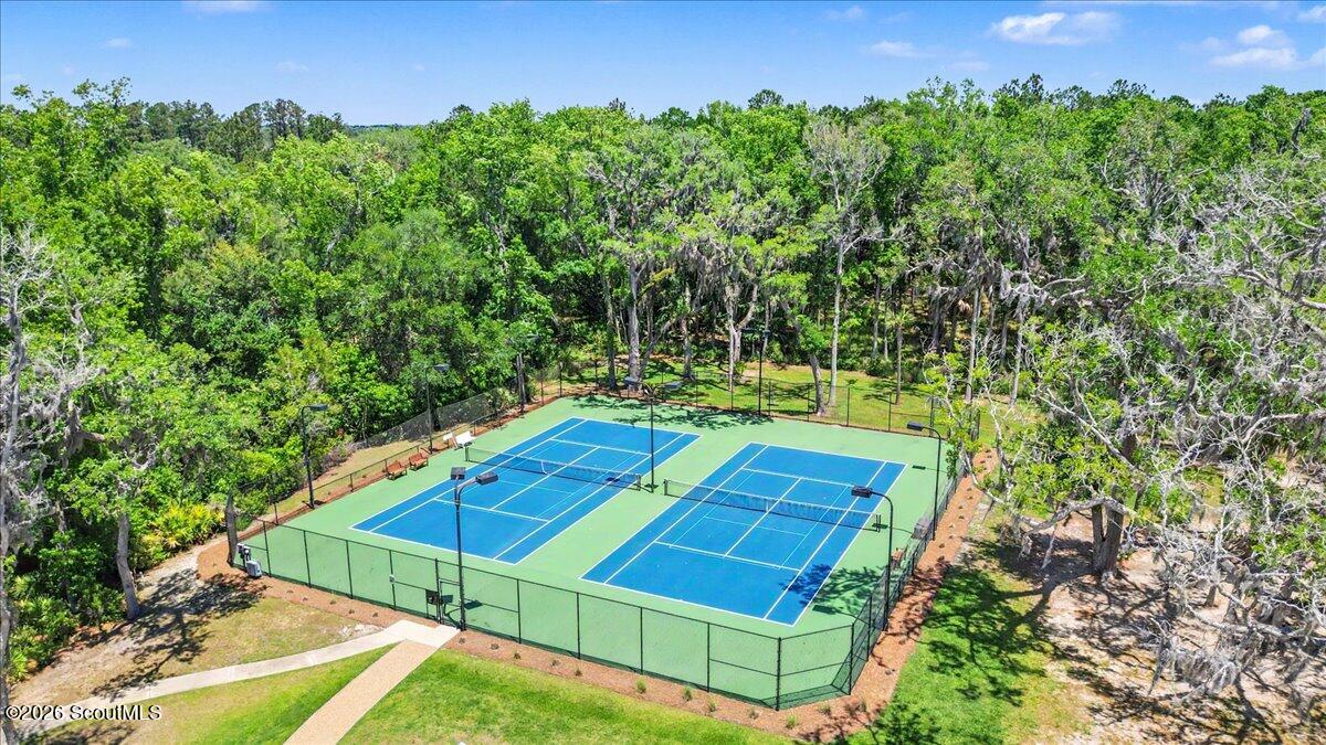 Two blue tennis courts sit amid dense green trees, with a paved walkway and lawn nearby.