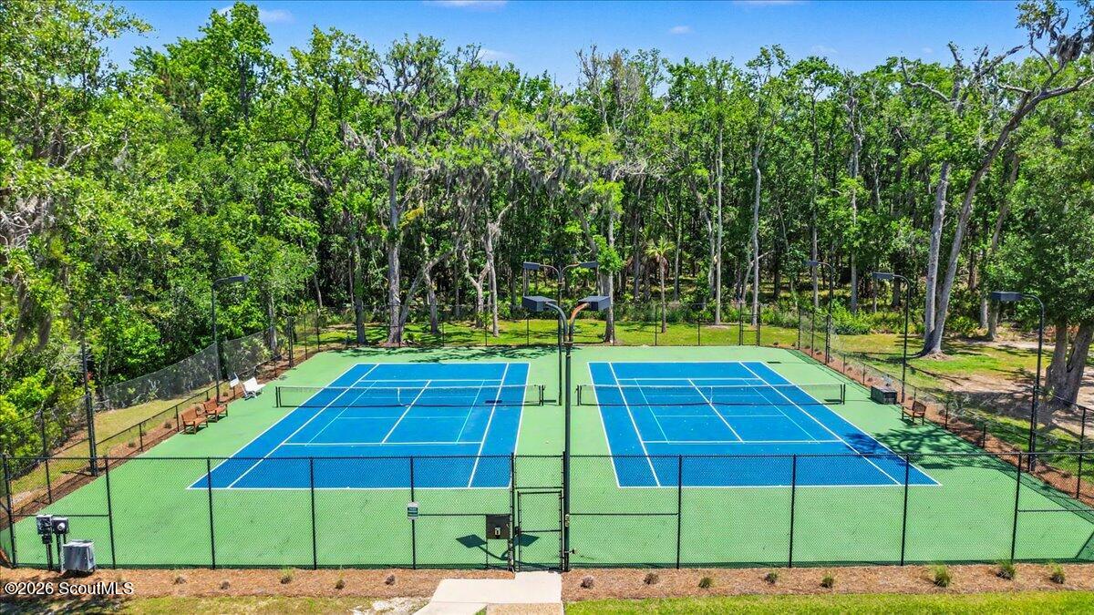 Two fenced blue tennis courts sit among dense trees, with benches and court lights.