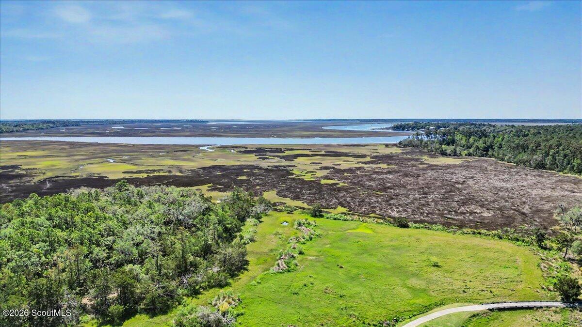 Aerial view of green land beside wooded marshland, coastal waterways, and a curving paved road.