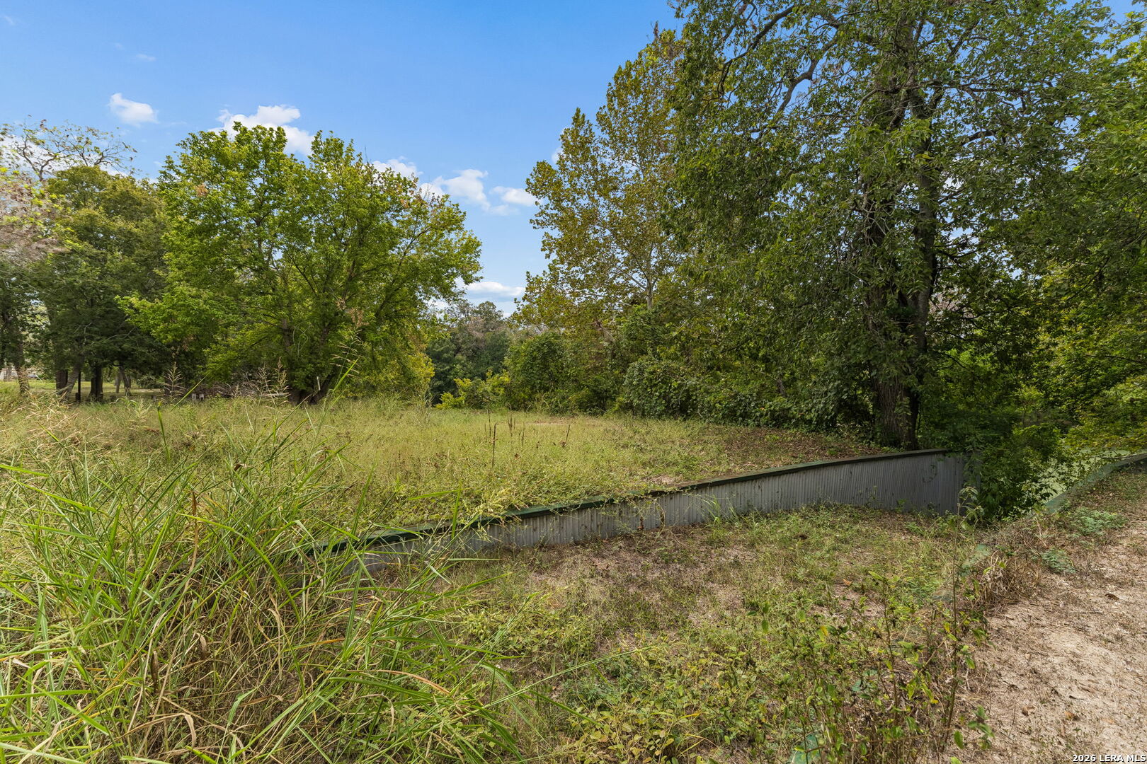 Wooded grassy lot with a clearing, mature trees, and a low retaining wall.
