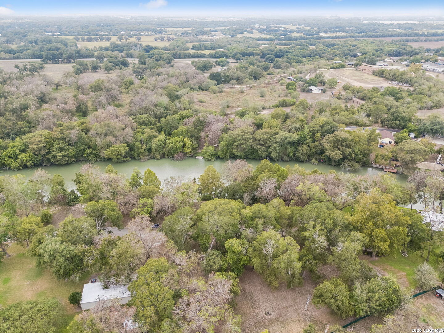 Aerial view of a wooded property beside a river, with a dock and homes along the opposite bank.