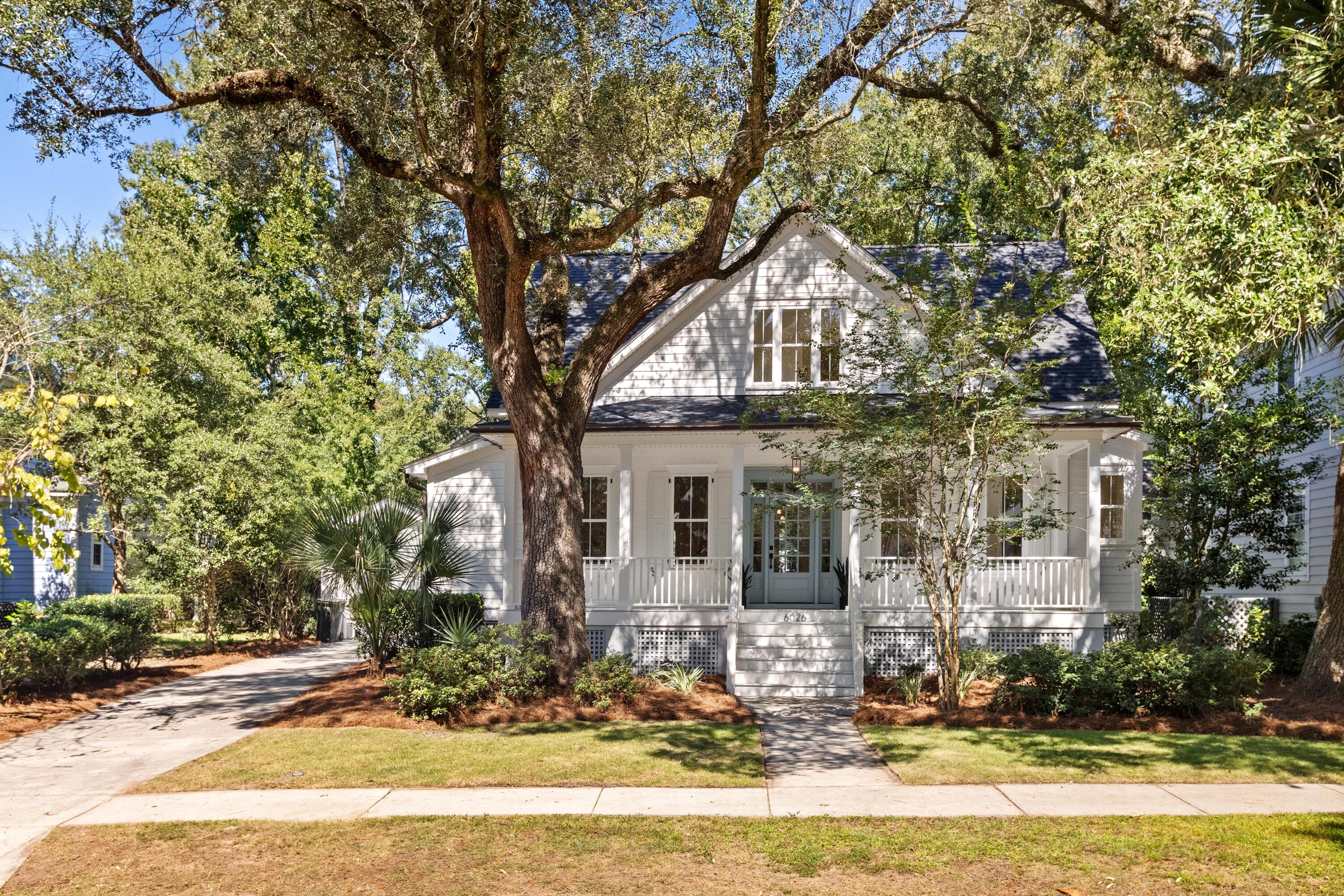 White two-story home with a front porch beneath a large oak tree, surrounded by mature landscaping.
