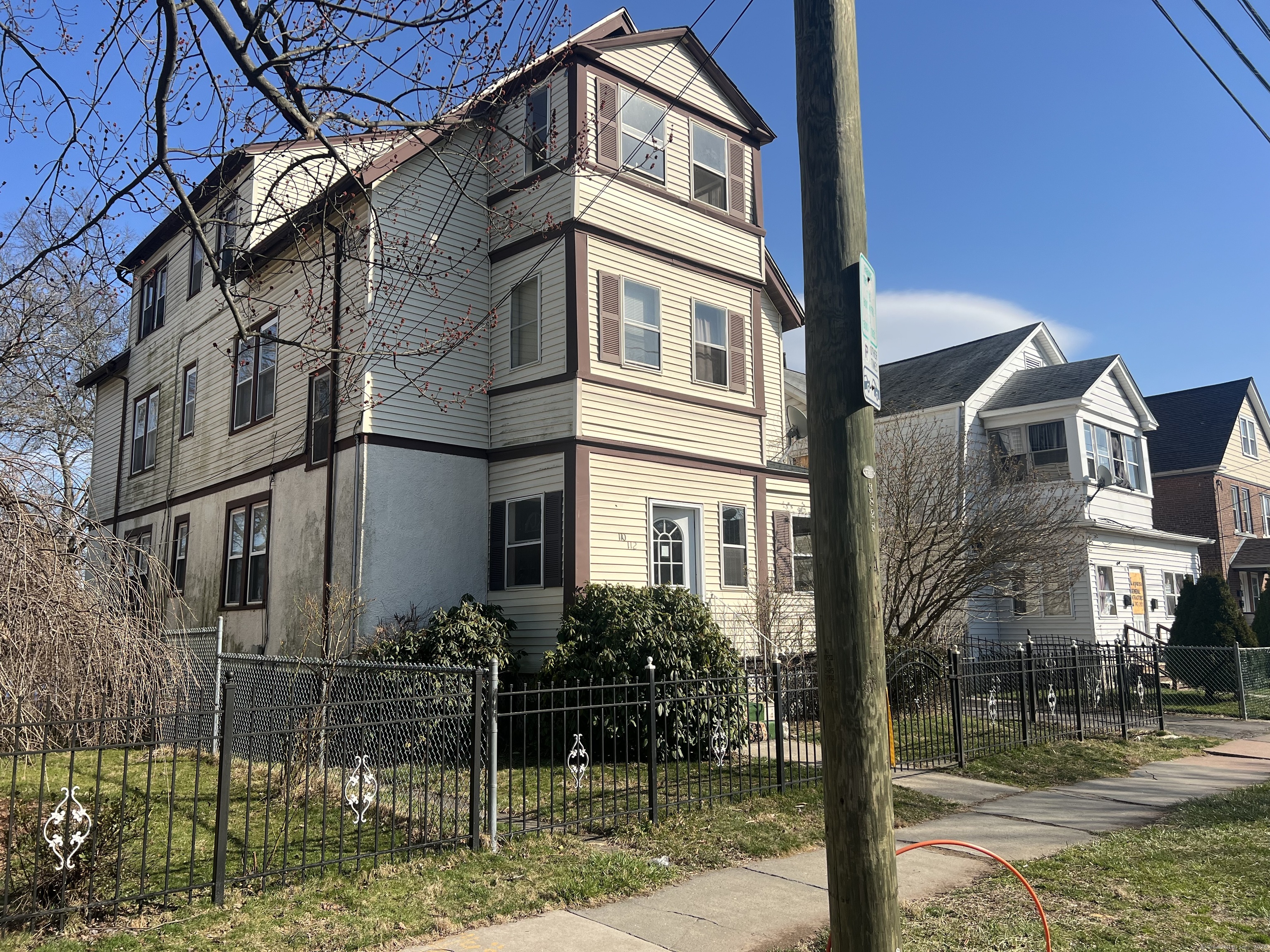 Tall cream-sided house with brown trim behind a metal fence, partly obscured by a utility pole.