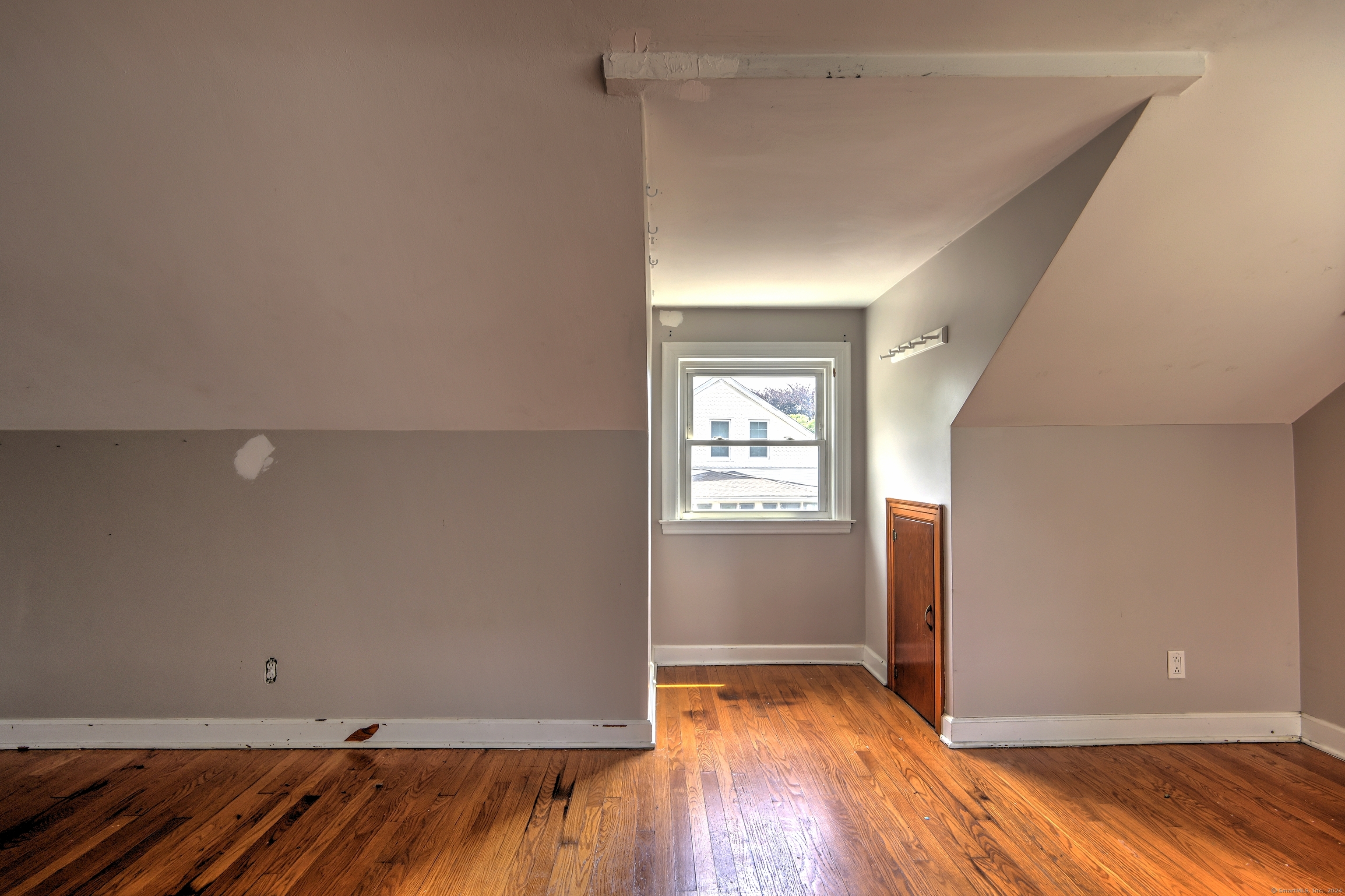 Empty upper-floor room with sloped ceilings, hardwood floors, a window, and a narrow wooden cabinet.