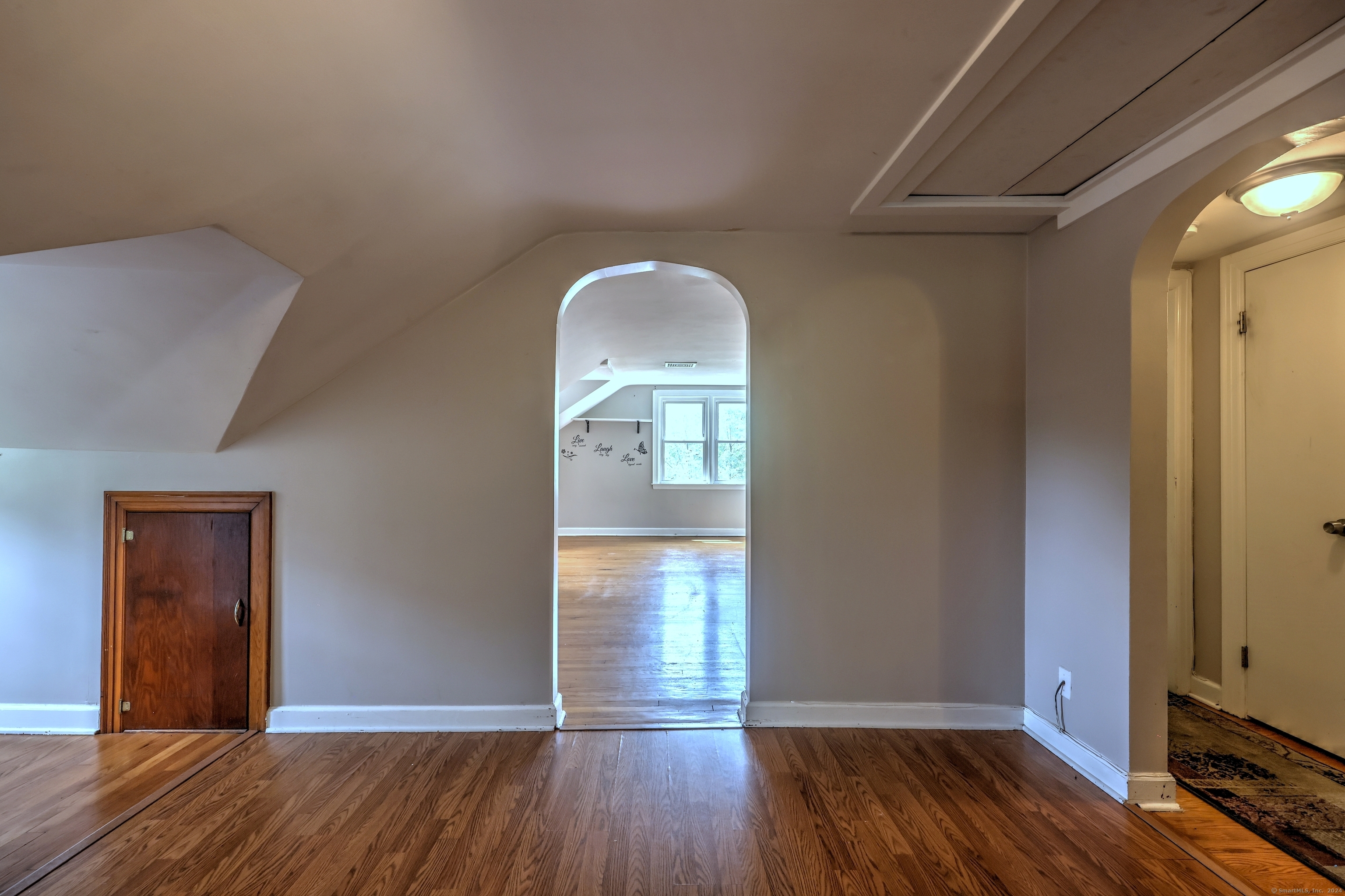 Empty hardwood-floored room with a sloped ceiling and arched doorway to a bright room.