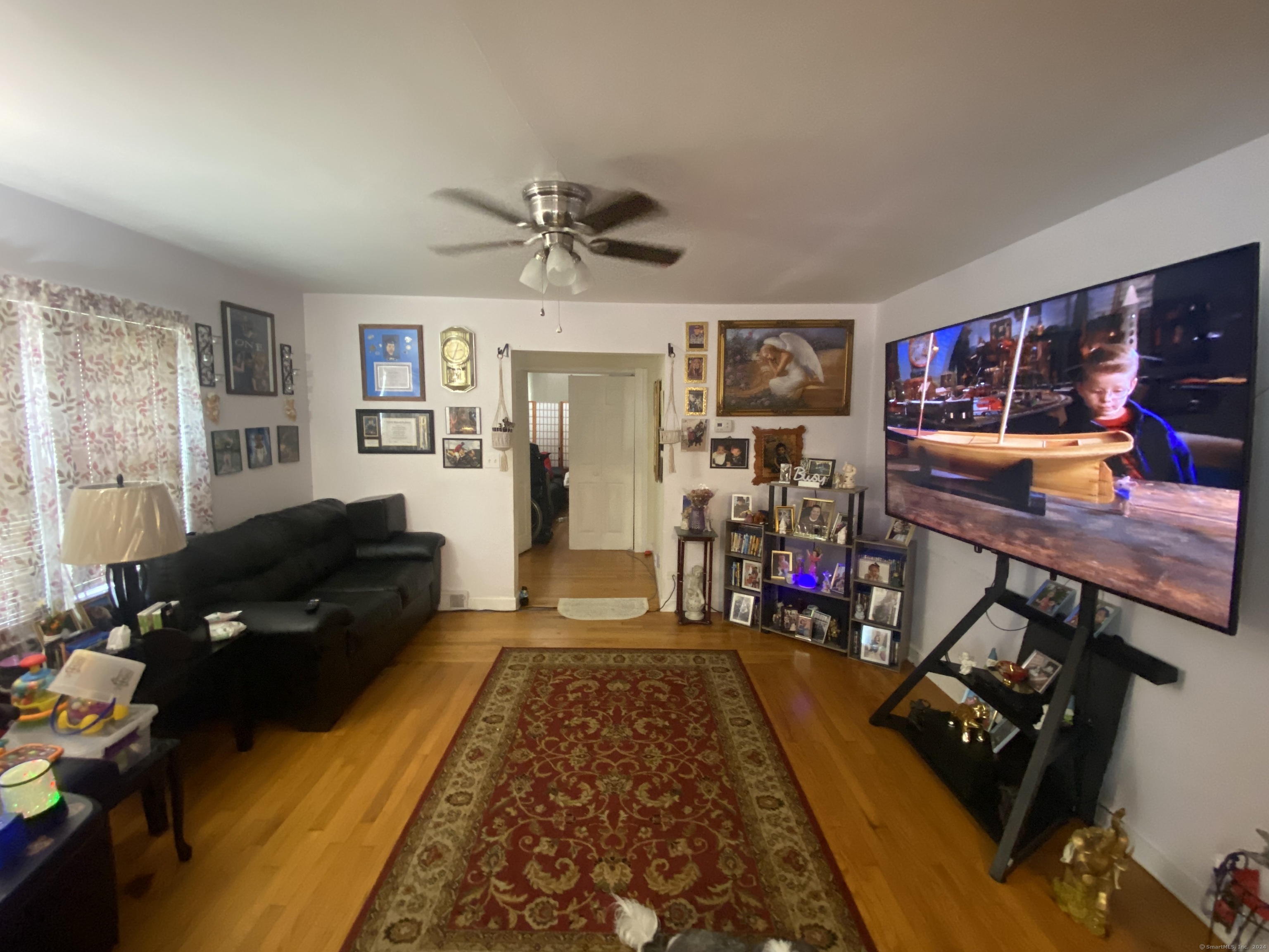 Living room with hardwood floors, a black sectional, red patterned rug, and large television.
