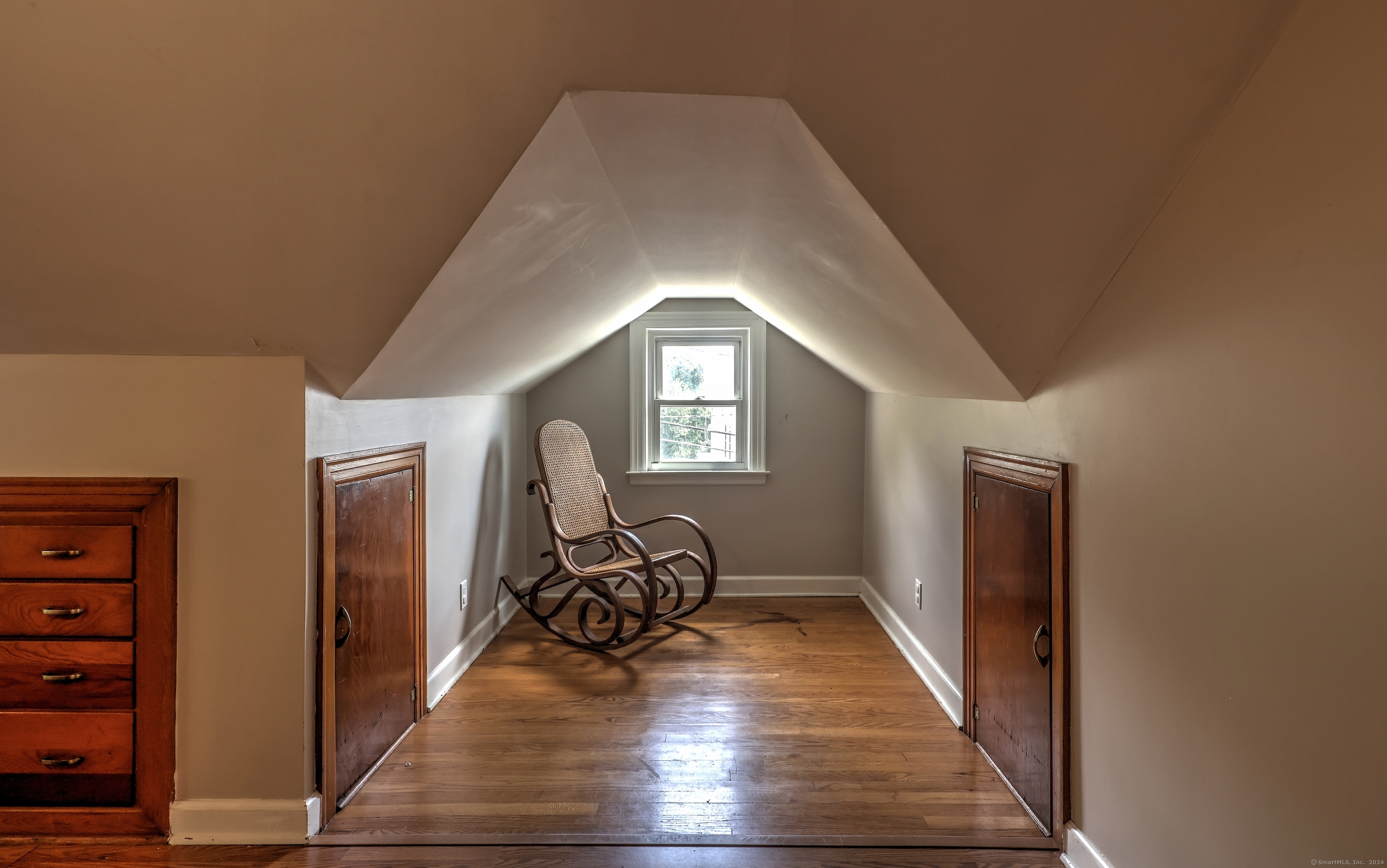 Furnished attic with a vintage rocking chair, hardwood floor, and window beneath a steeply pitched ceiling.