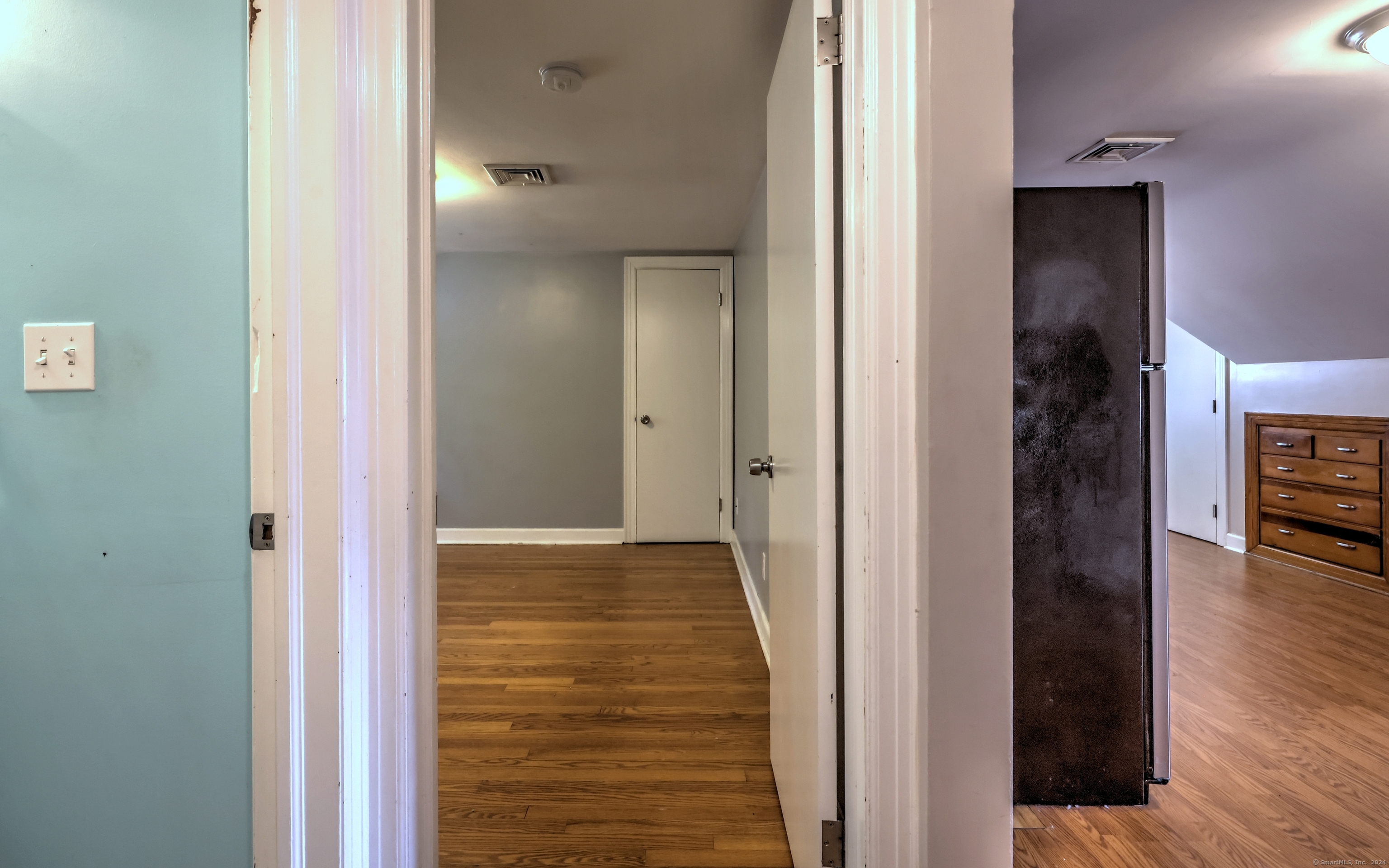 Hallway with hardwood floors, a closed door, and a view of a refrigerator and dresser in the adjoining room.