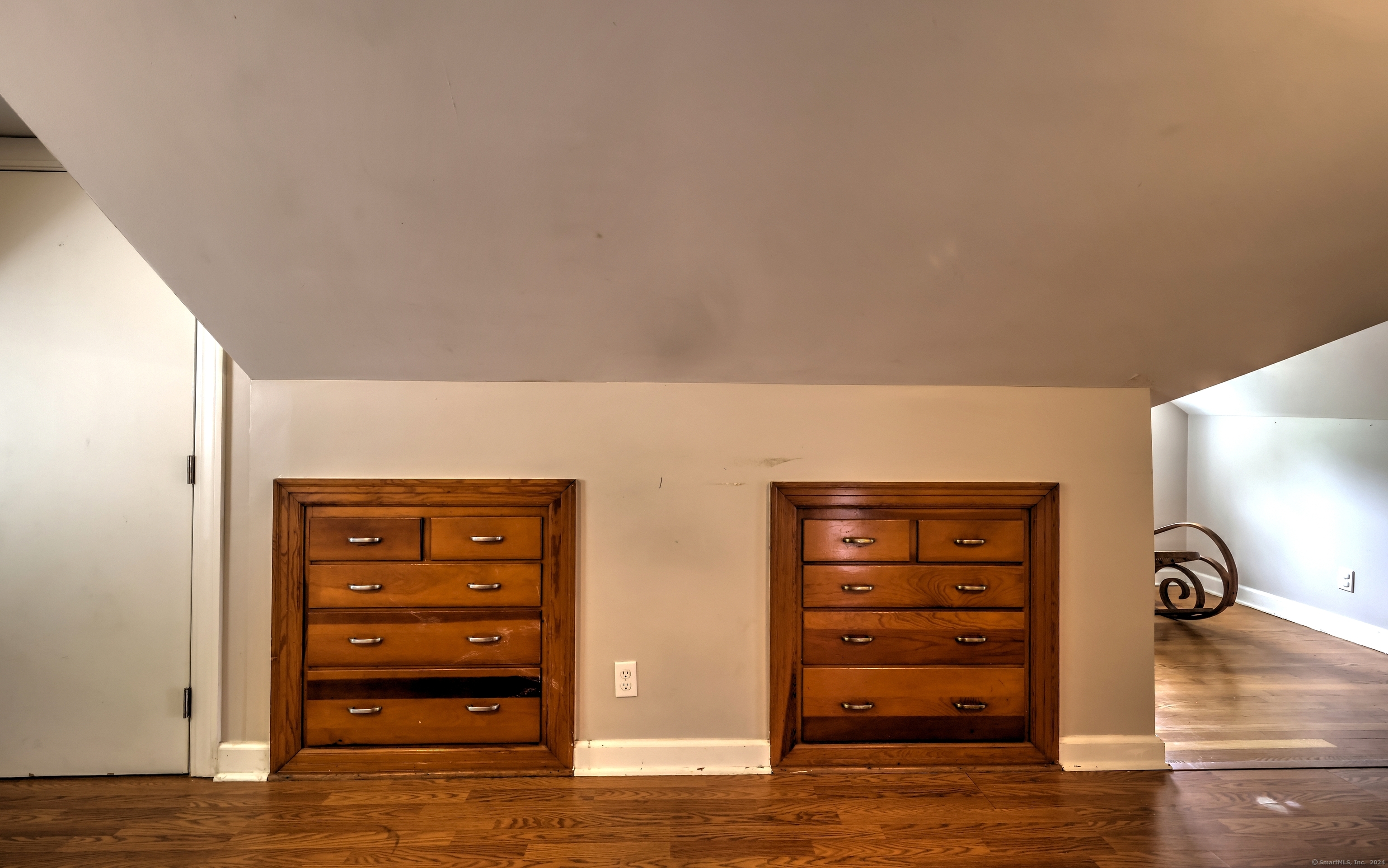 Built-in wooden drawers beneath a sloped ceiling, with hardwood floors and a rocking chair nearby.