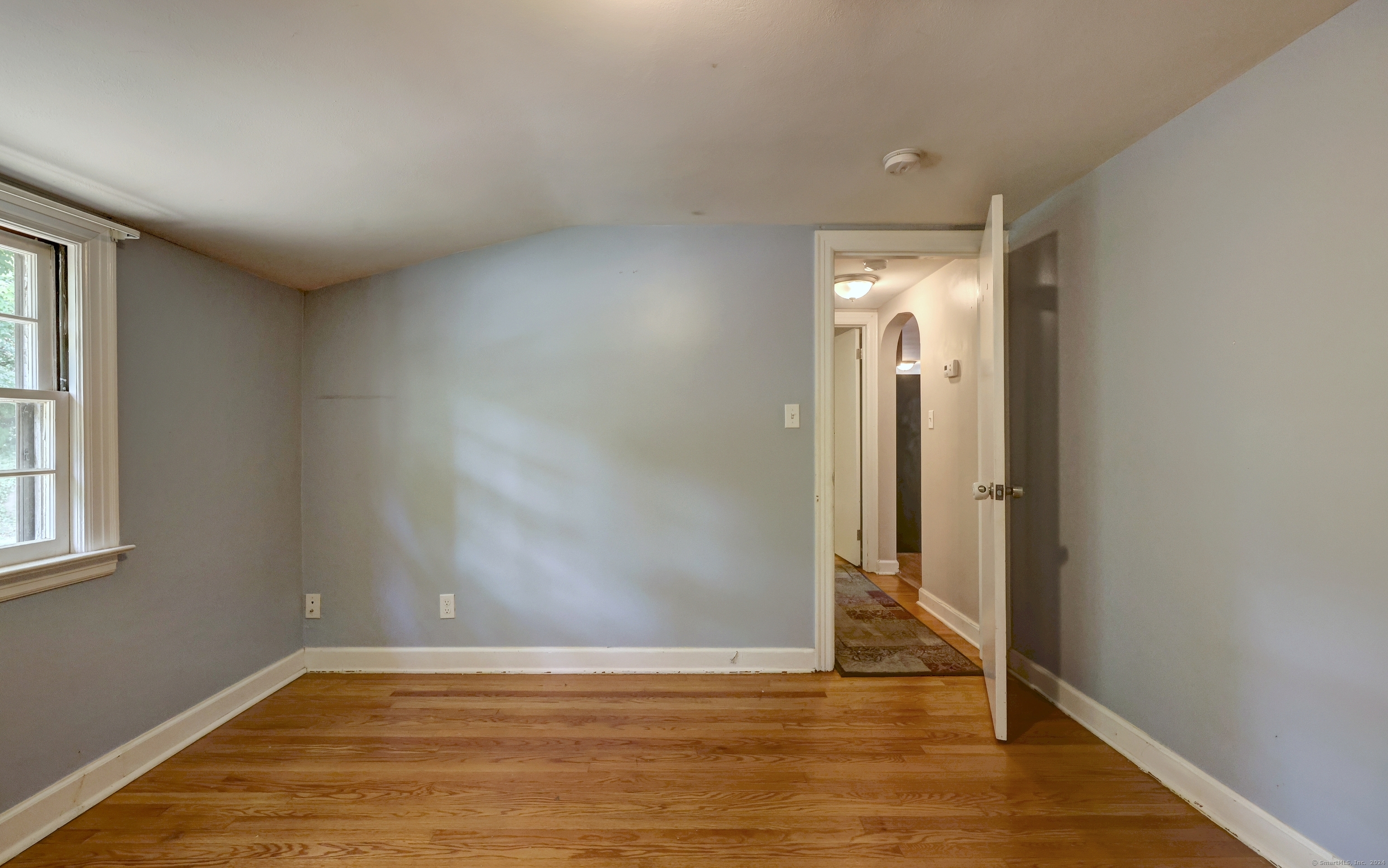 Empty room with hardwood floors, a window, and an open doorway to the hallway.
