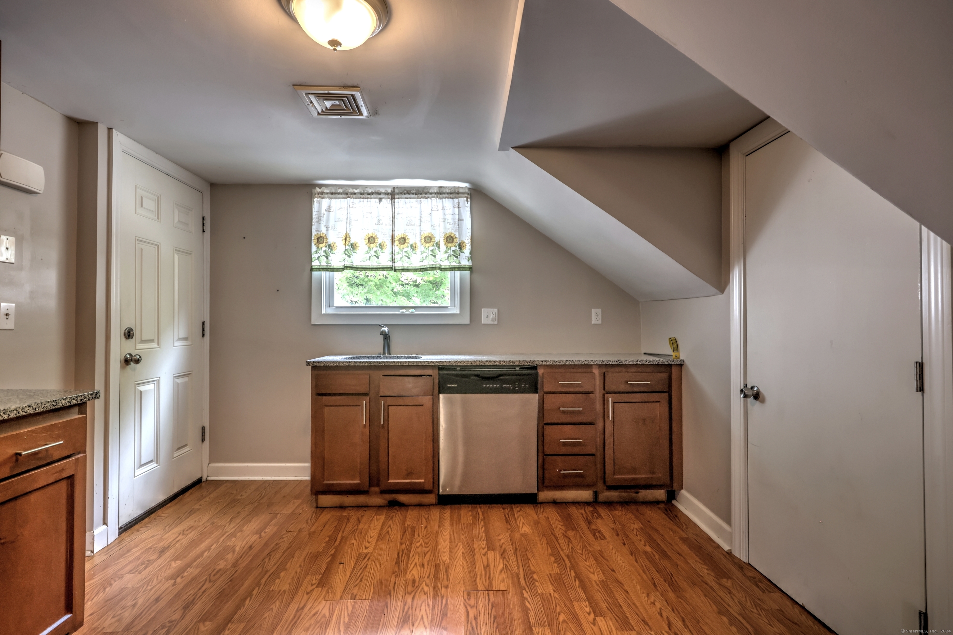 Kitchen with brown cabinets, granite counters, stainless dishwasher, and sunflower curtains beneath a sloped ceiling.