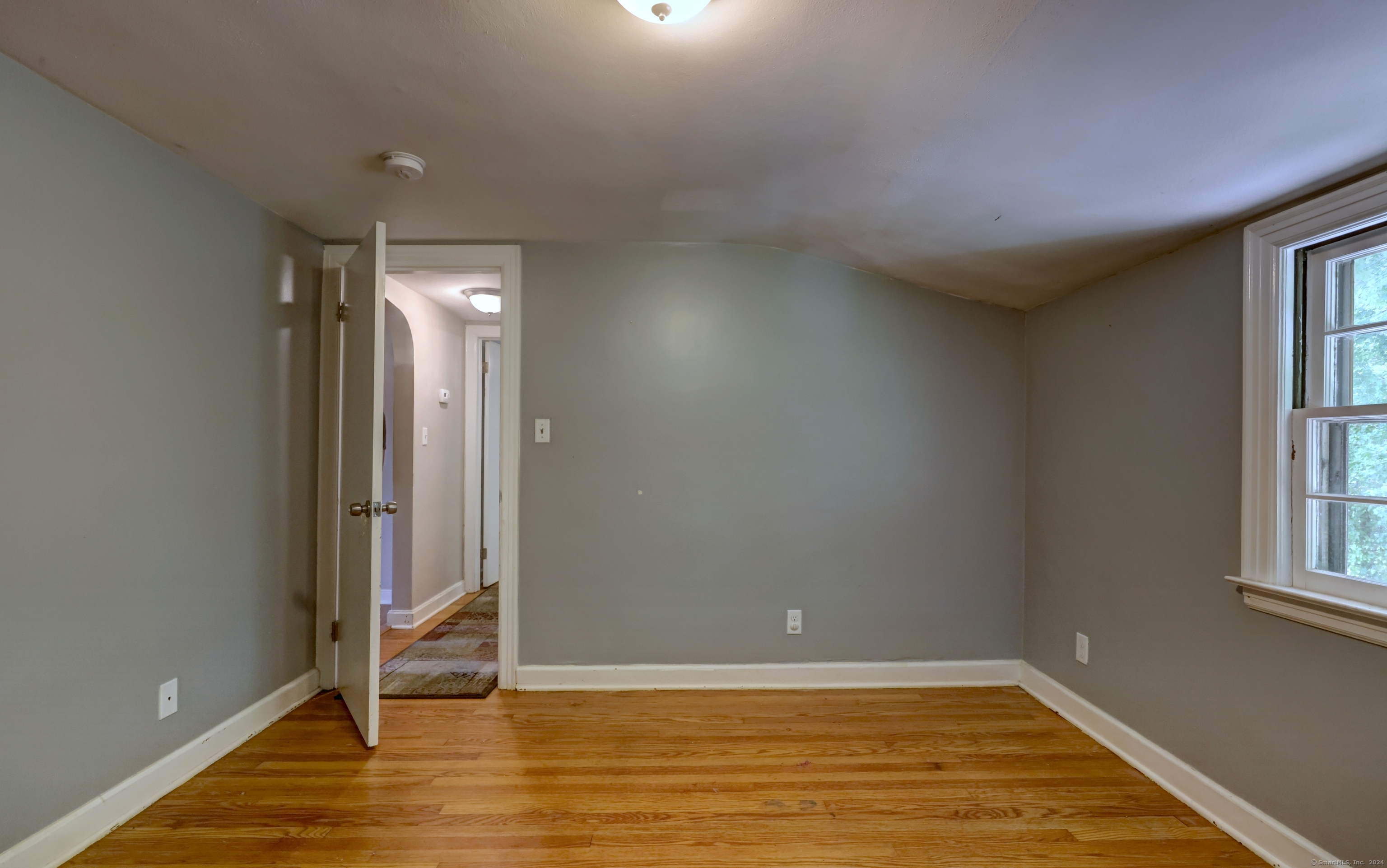 Empty gray-walled room with hardwood floors and a window beside an open doorway.