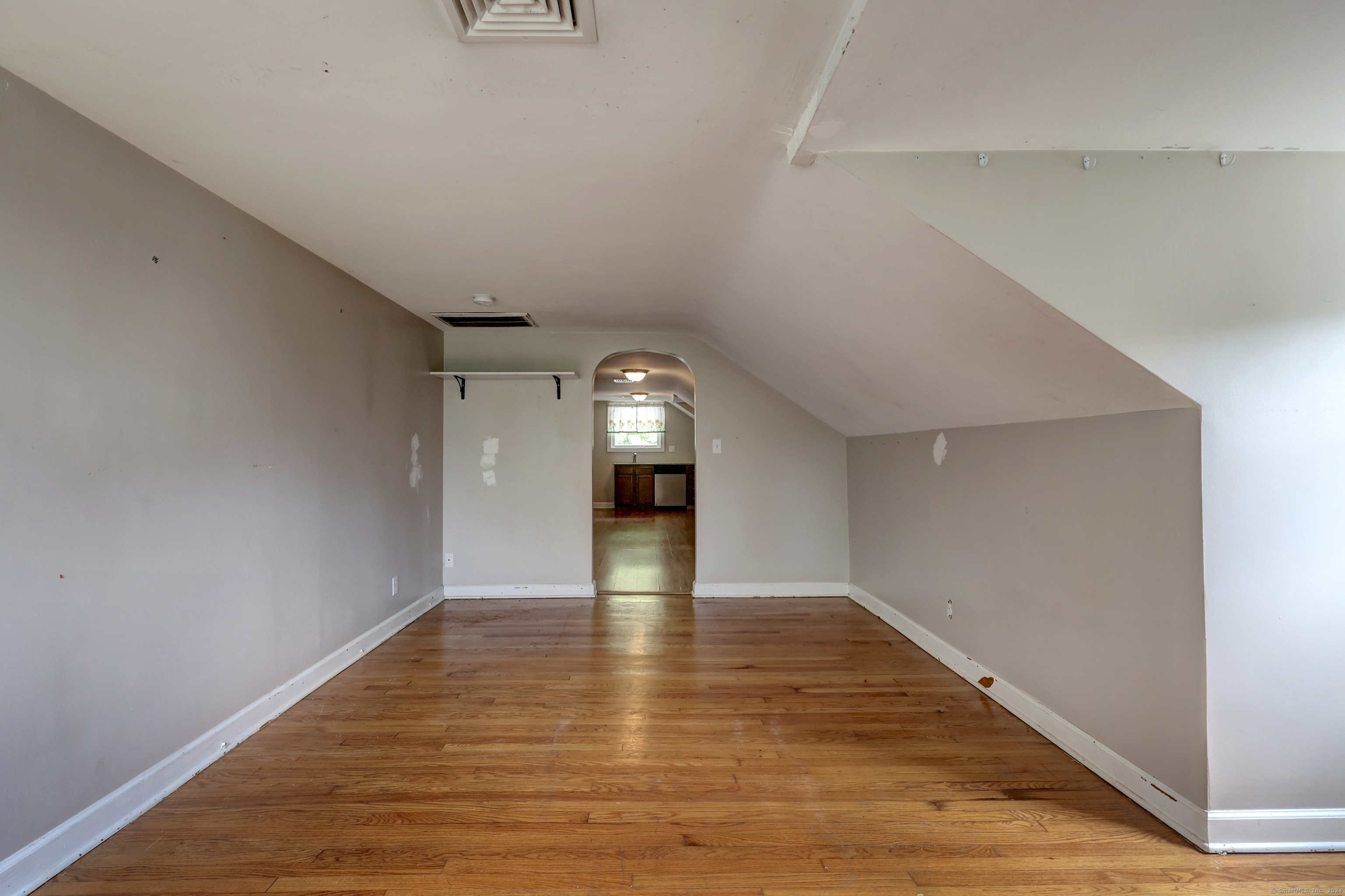 Vacant room with sloped ceilings and hardwood floors, looking through an archway toward the kitchen.