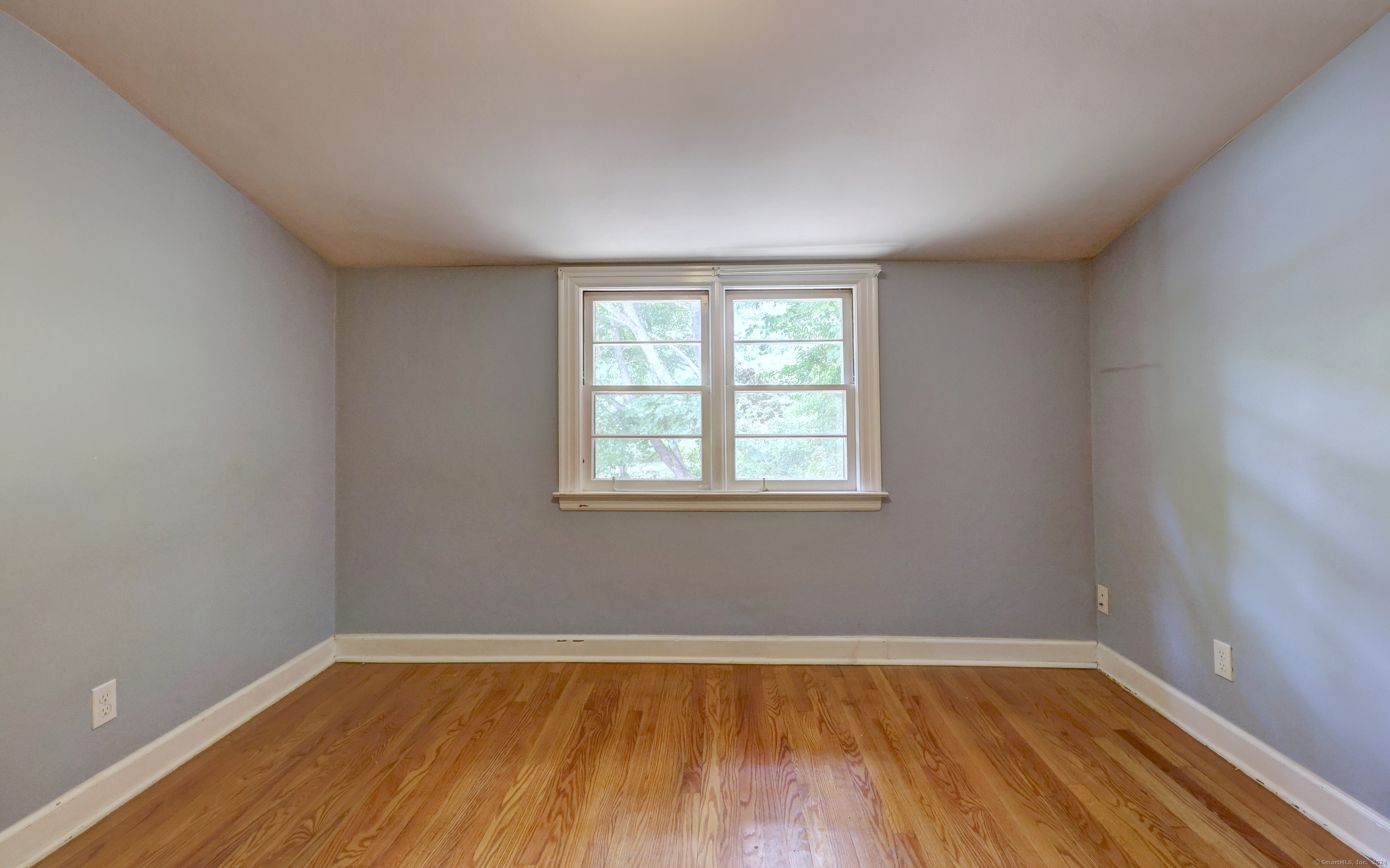 Empty room with polished hardwood floors, gray walls, and a double window overlooking trees.