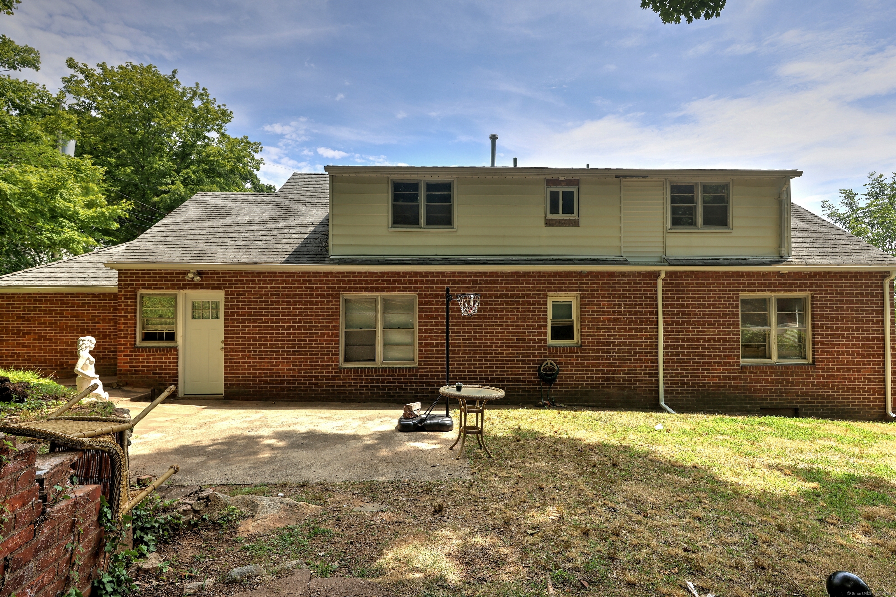 House rear with brick lower level, pale upper siding, patio table, and basketball hoop.