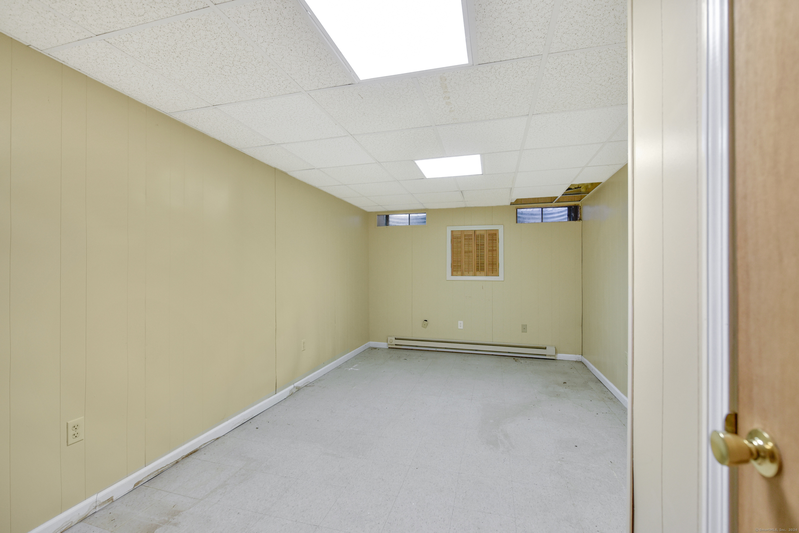 Empty pale yellow room with gray flooring, fluorescent ceiling lights, high windows, and a baseboard heater.