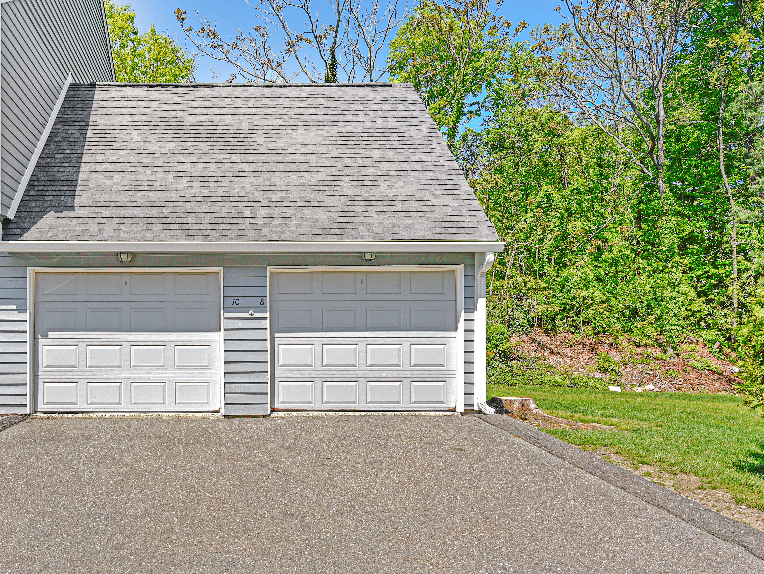 Gray-sided home with two white garage doors, a broad asphalt driveway, and trees beside the lawn.
