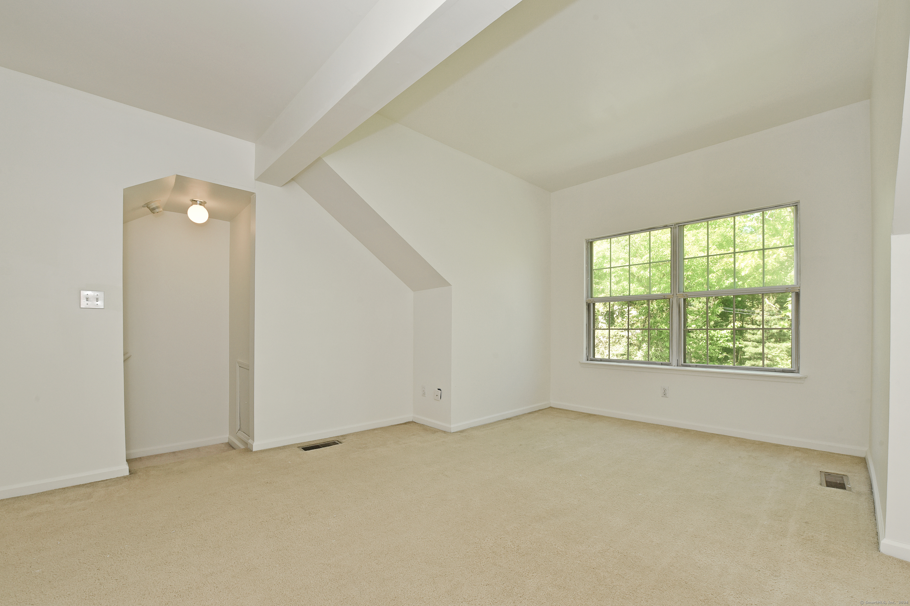 Empty carpeted room with an angled ceiling and large window overlooking trees.
