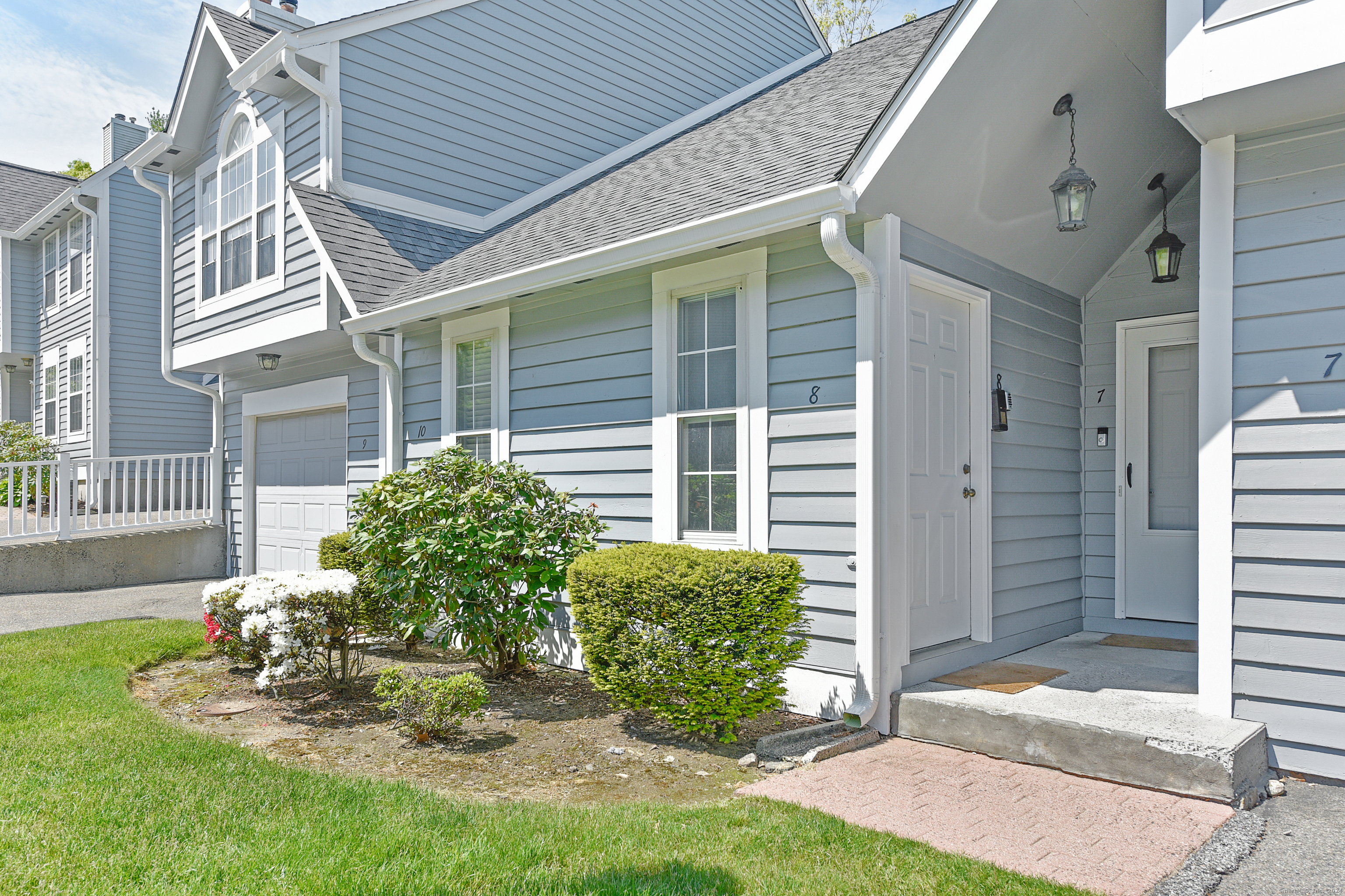 Blue-gray attached home with white trim, a covered entrance, and shrubs along the front.