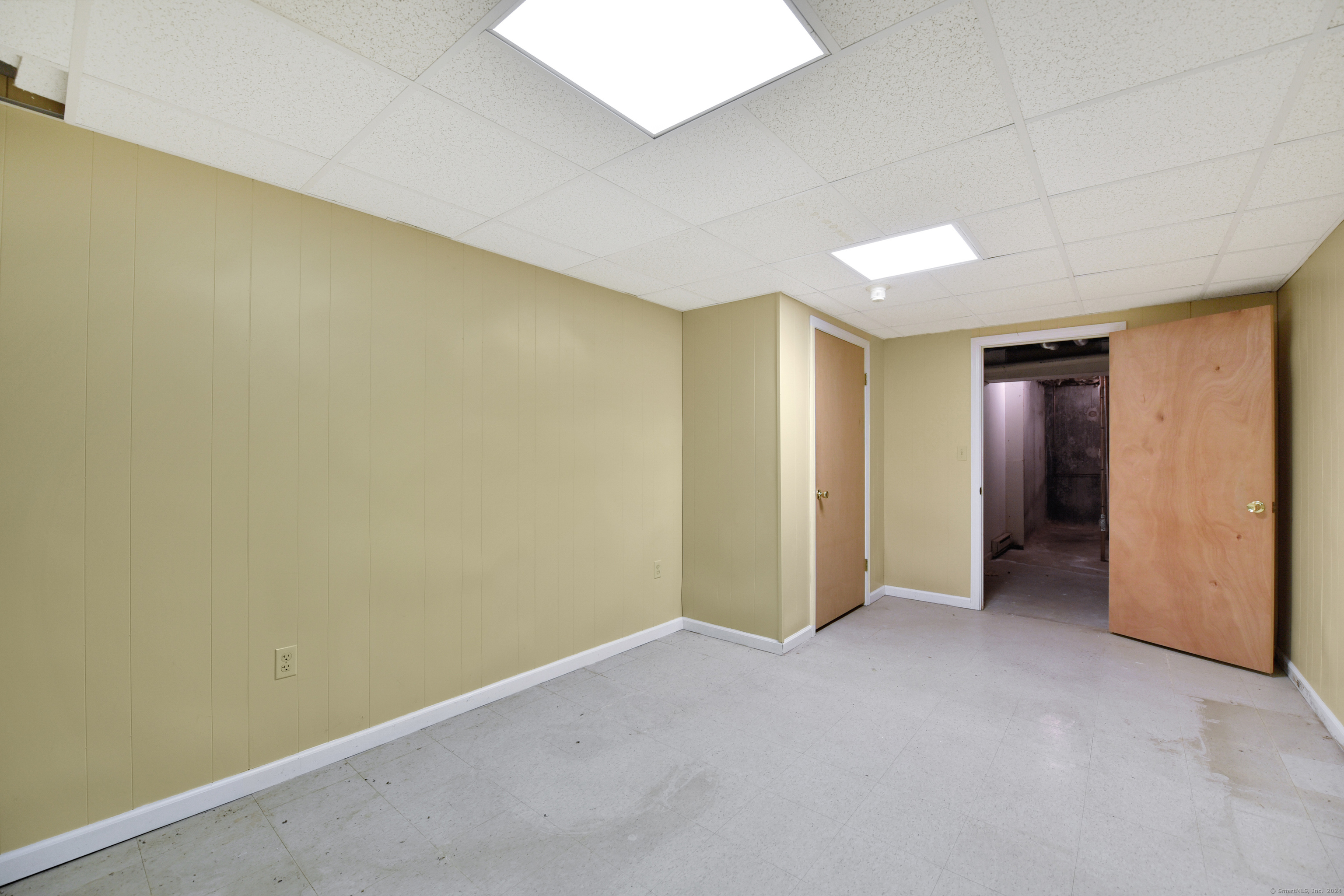 Empty basement room with pale paneled walls, tiled floor, and an open door to an unfinished area.
