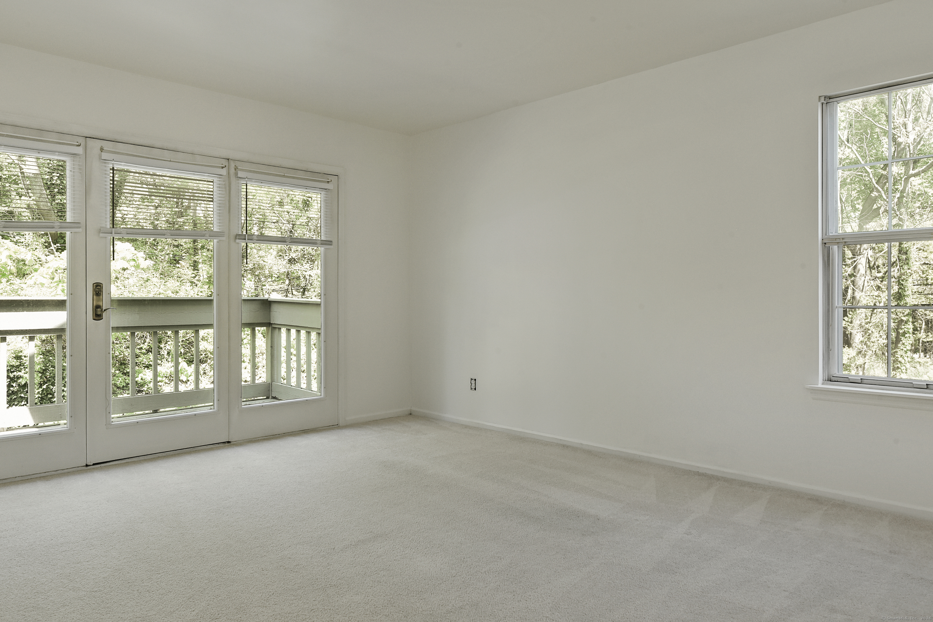 Empty carpeted room with glass balcony doors and a window overlooking trees.
