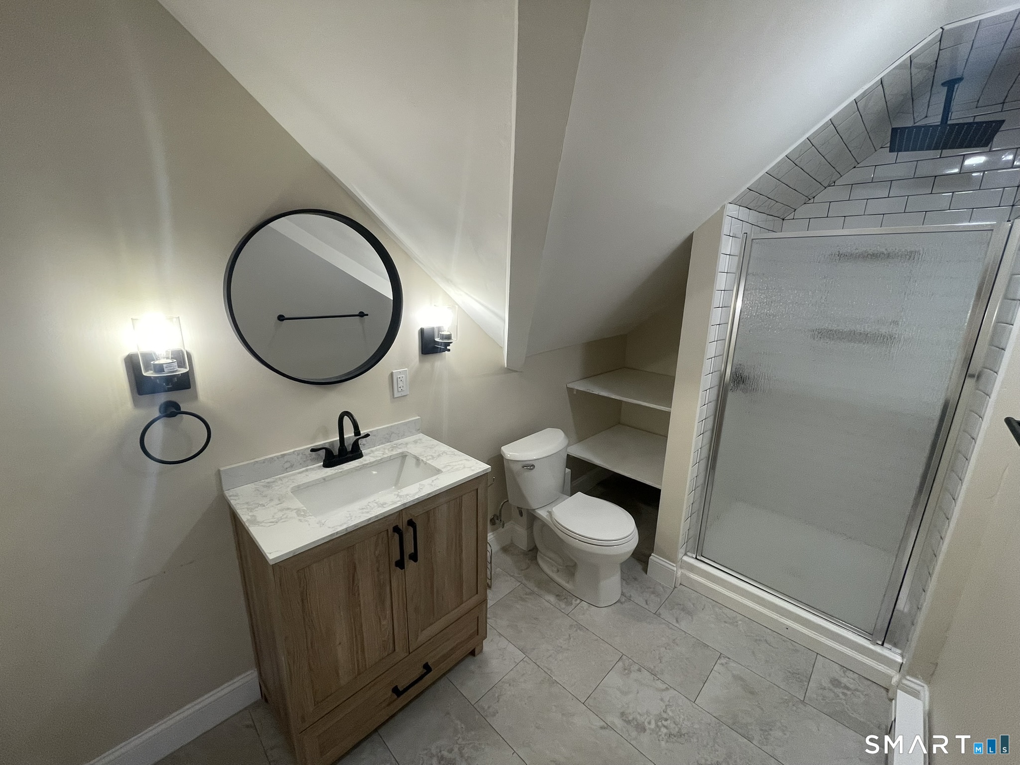 Bathroom with a wood vanity, round mirror, built-in shelves, and tiled shower.
