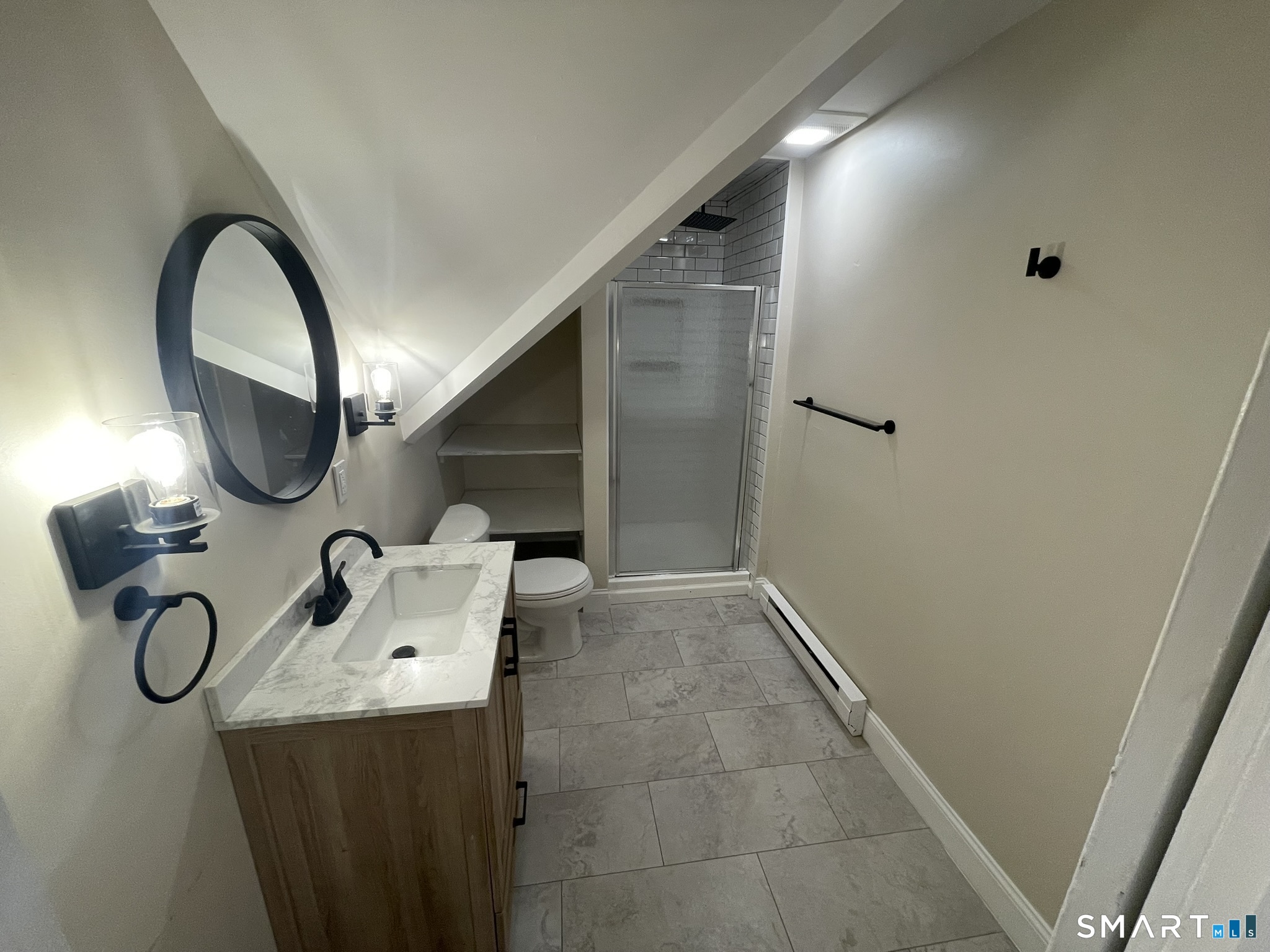 Bathroom with a wood vanity, oval mirror, and glass shower beneath a sloped ceiling.