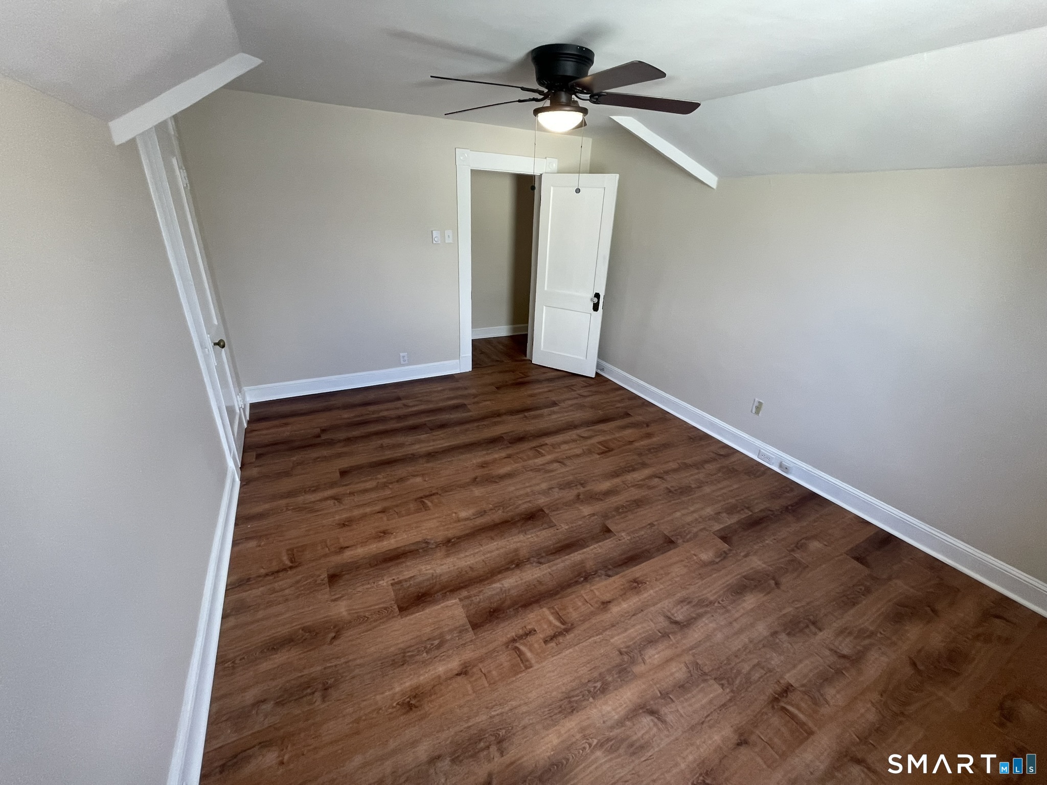 Empty attic bedroom with sloped ceilings, wood-look flooring, and a ceiling fan.