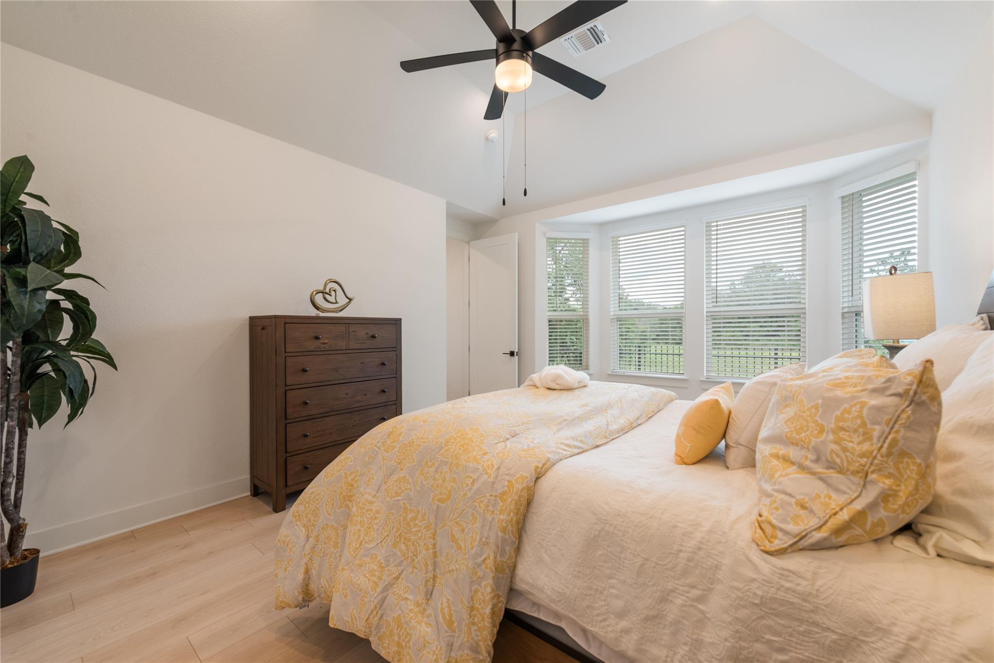 Bedroom with a white bed, yellow floral bedding, dark wood dresser, and bright bay windows.